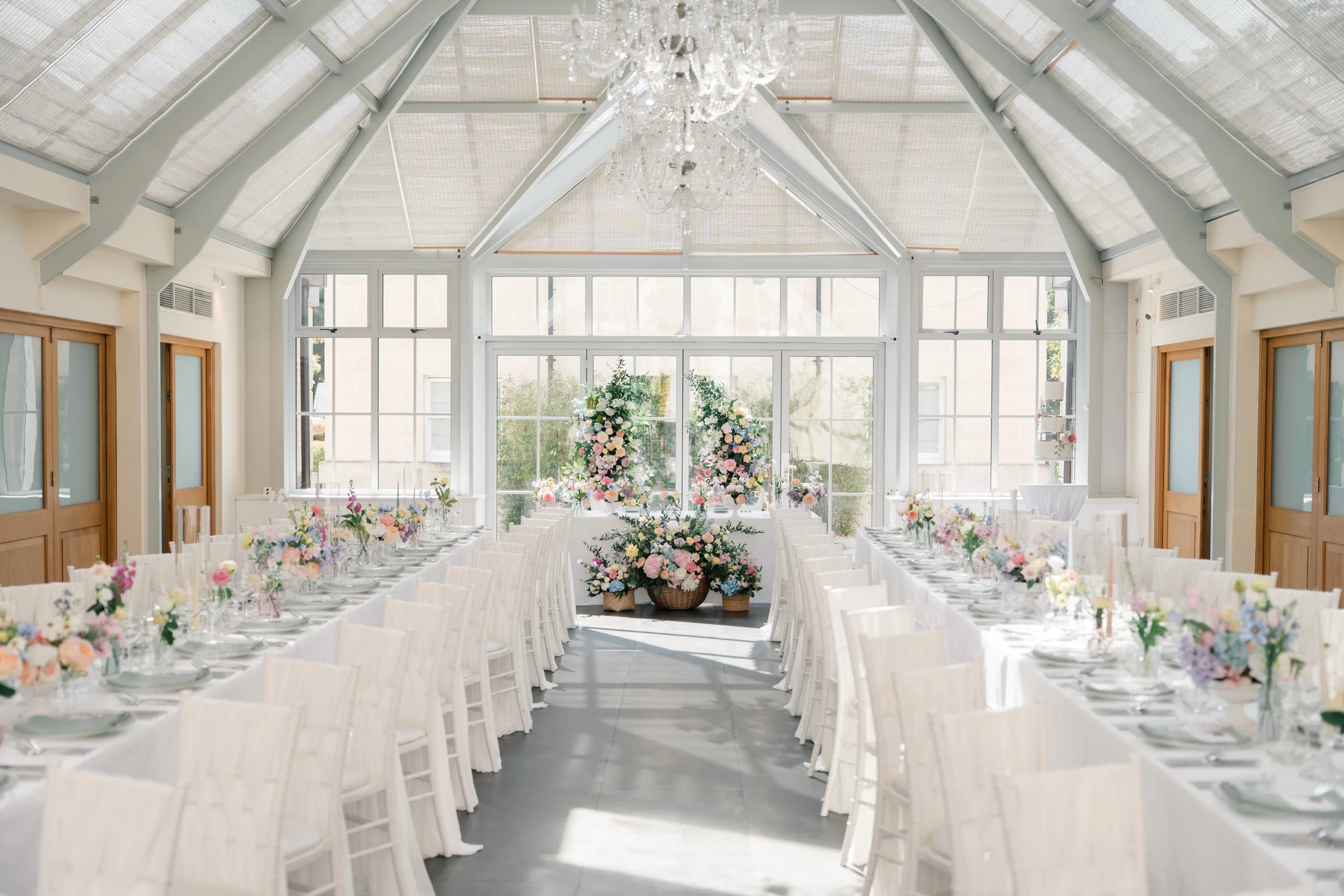 Elegant wedding reception setup in a bright glass conservatory, with long white-draped tables decorated with pastel floral centerpieces and a chandelier hanging from the ceiling.