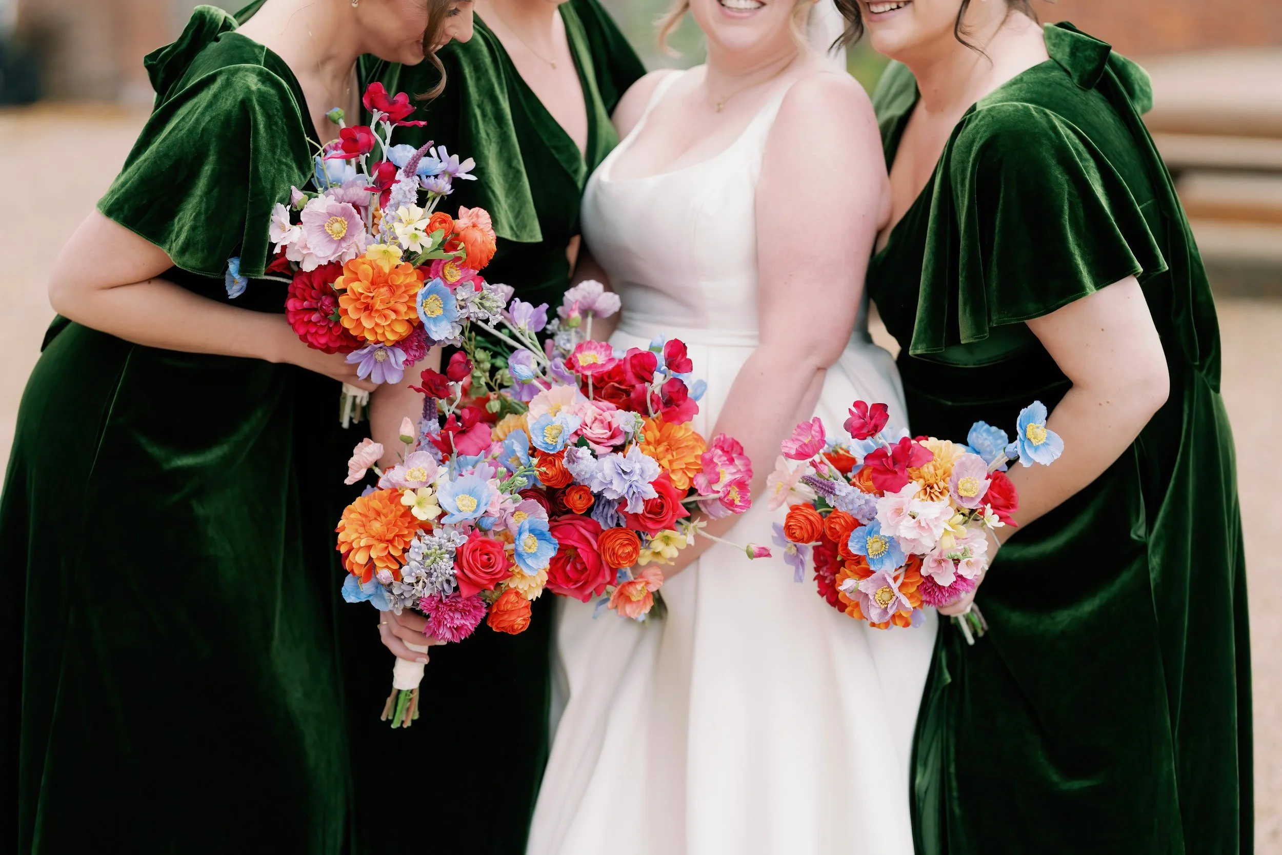 A bride in a white dress holding a colorful bouquet of flowers surrounded by three women in green velvet dresses, all holding smaller bouquets, outside during daytime.