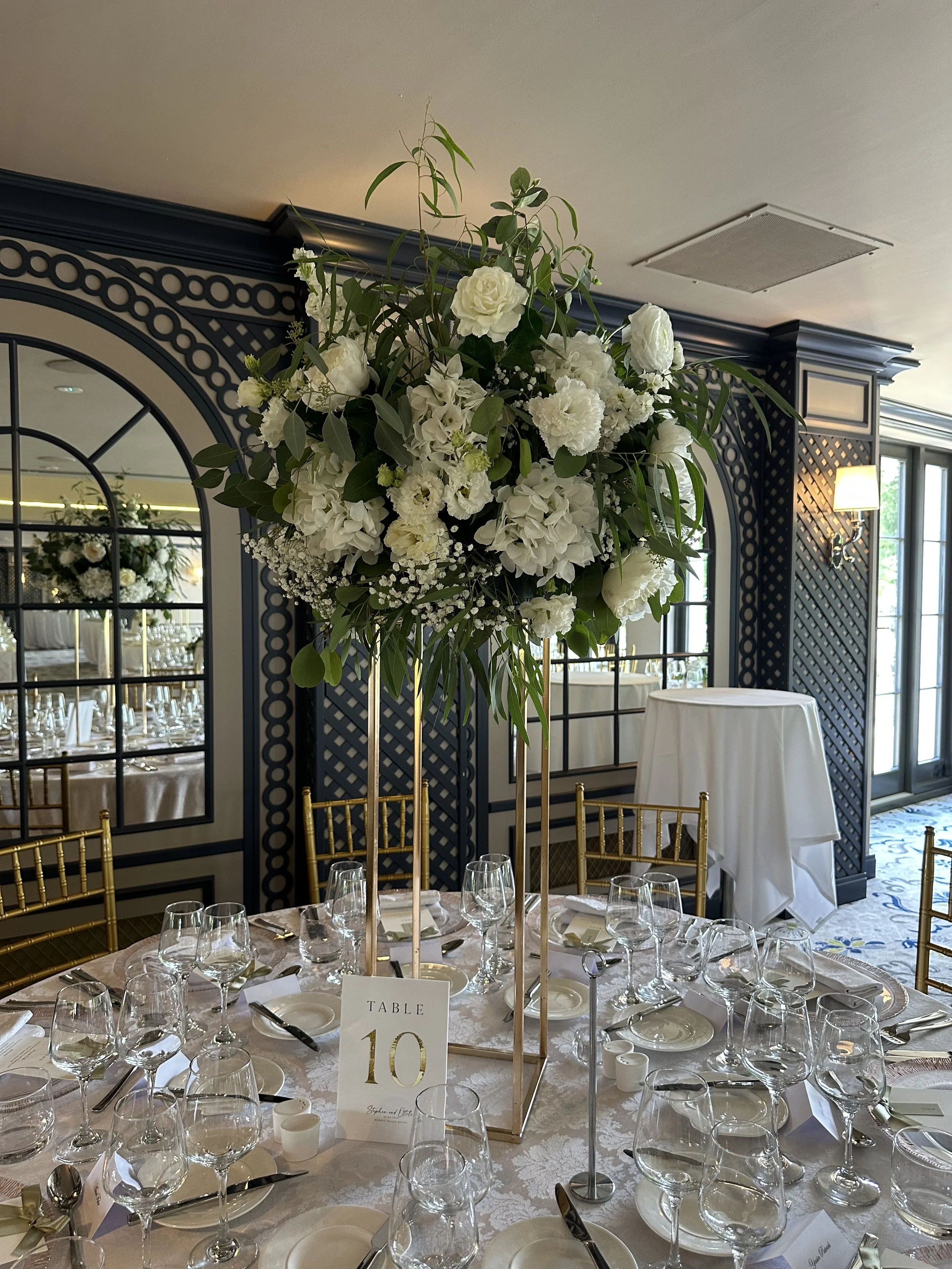 Elegant banquet table with a tall floral centerpiece of white flowers and greenery, set with multiple glasses, plates, and silverware in a decorated room with dark wall panels and large windows.
