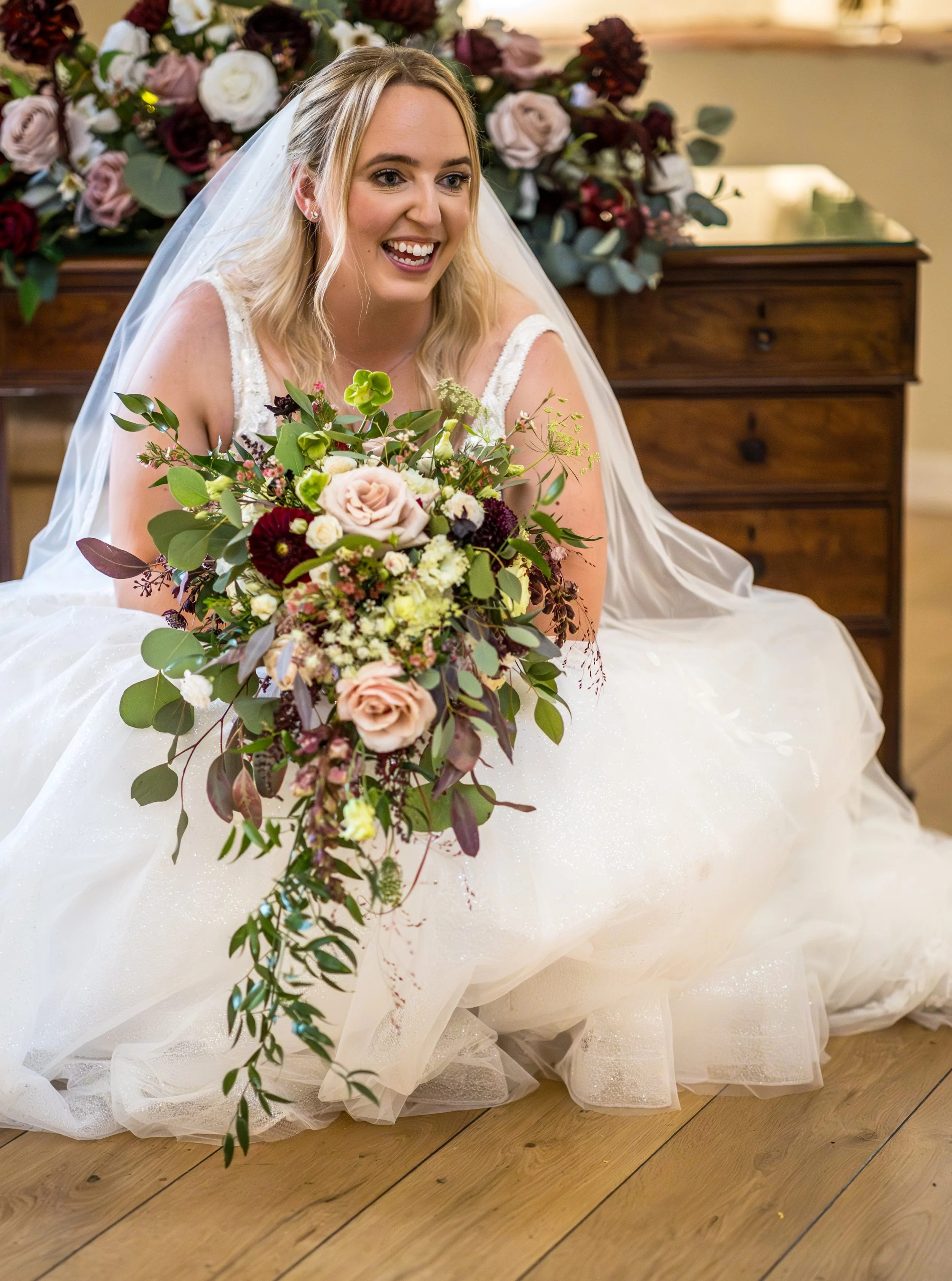 A smiling bride in a wedding dress holds a large bouquet of flowers, sitting on a wooden floor in front of a floral arrangement.
