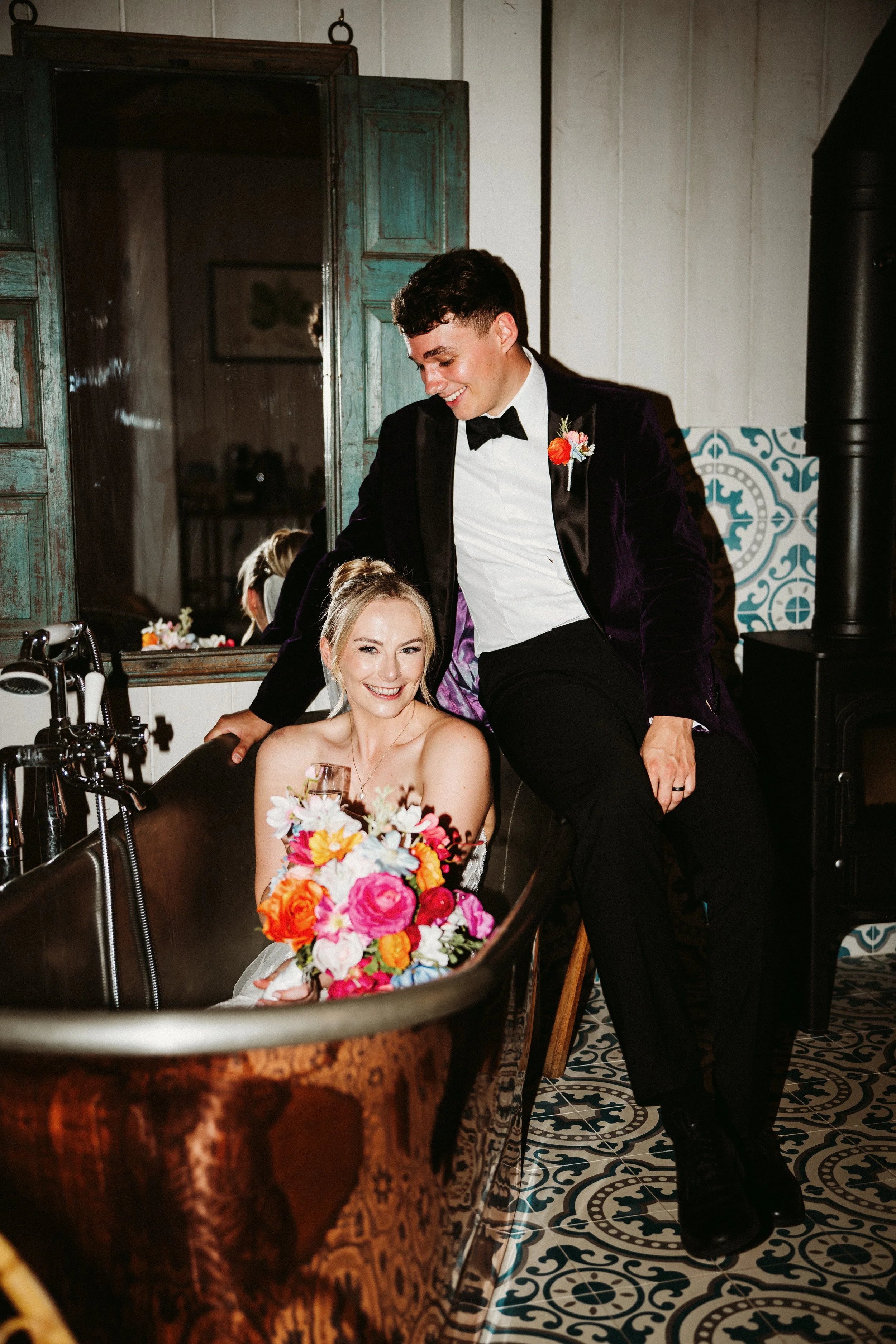 A bride sitting in a vintage copper bathtub holding a colorful bouquet, smiling at the camera, with a groom standing beside her leaning on the tub, both dressed in formal wedding attire, in a rustic indoor setting.