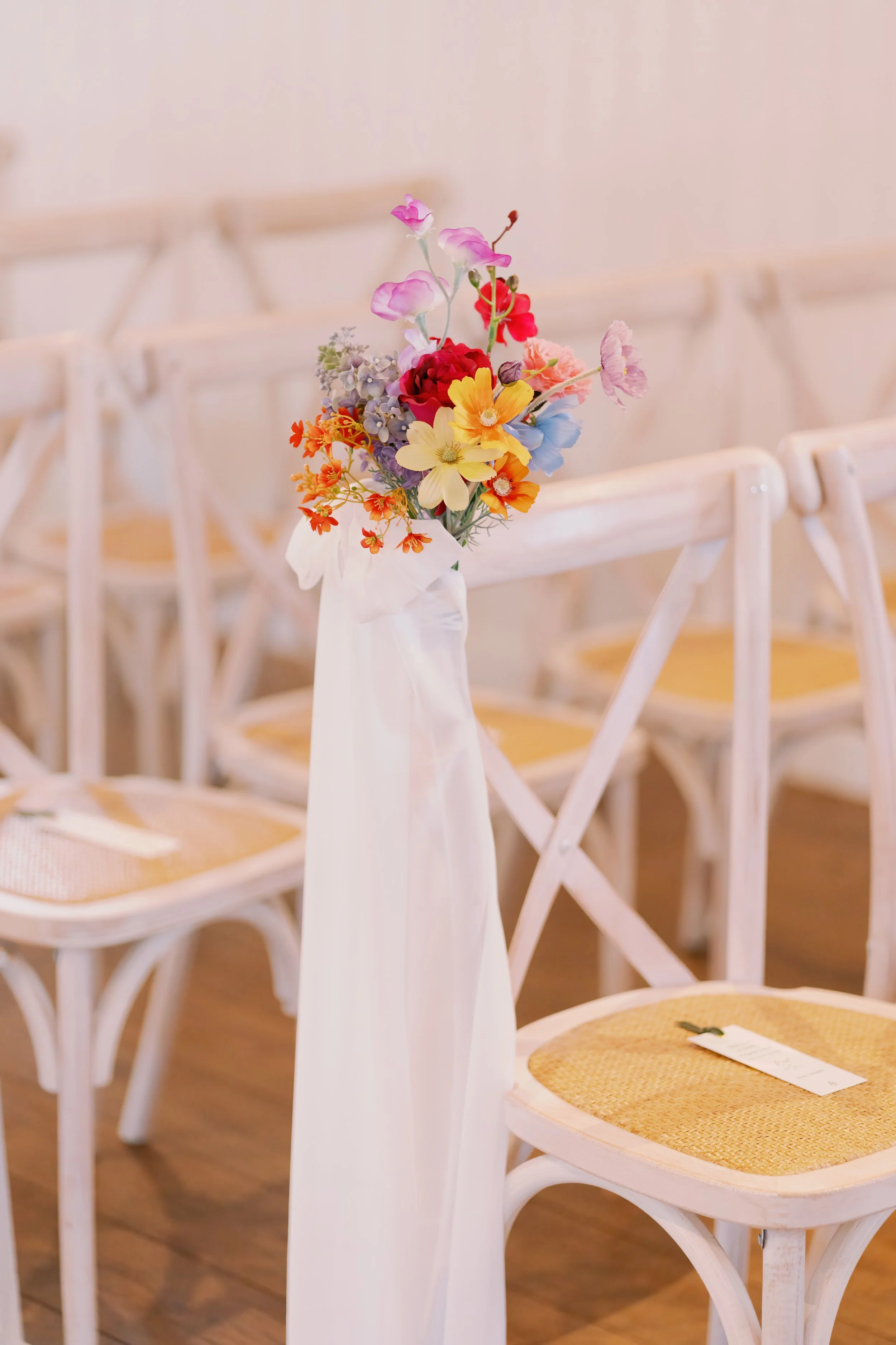 A white fabric tied to a chair back holding a colorful flower bouquet in a white vase, surrounded by empty white wooden chairs with woven seats.