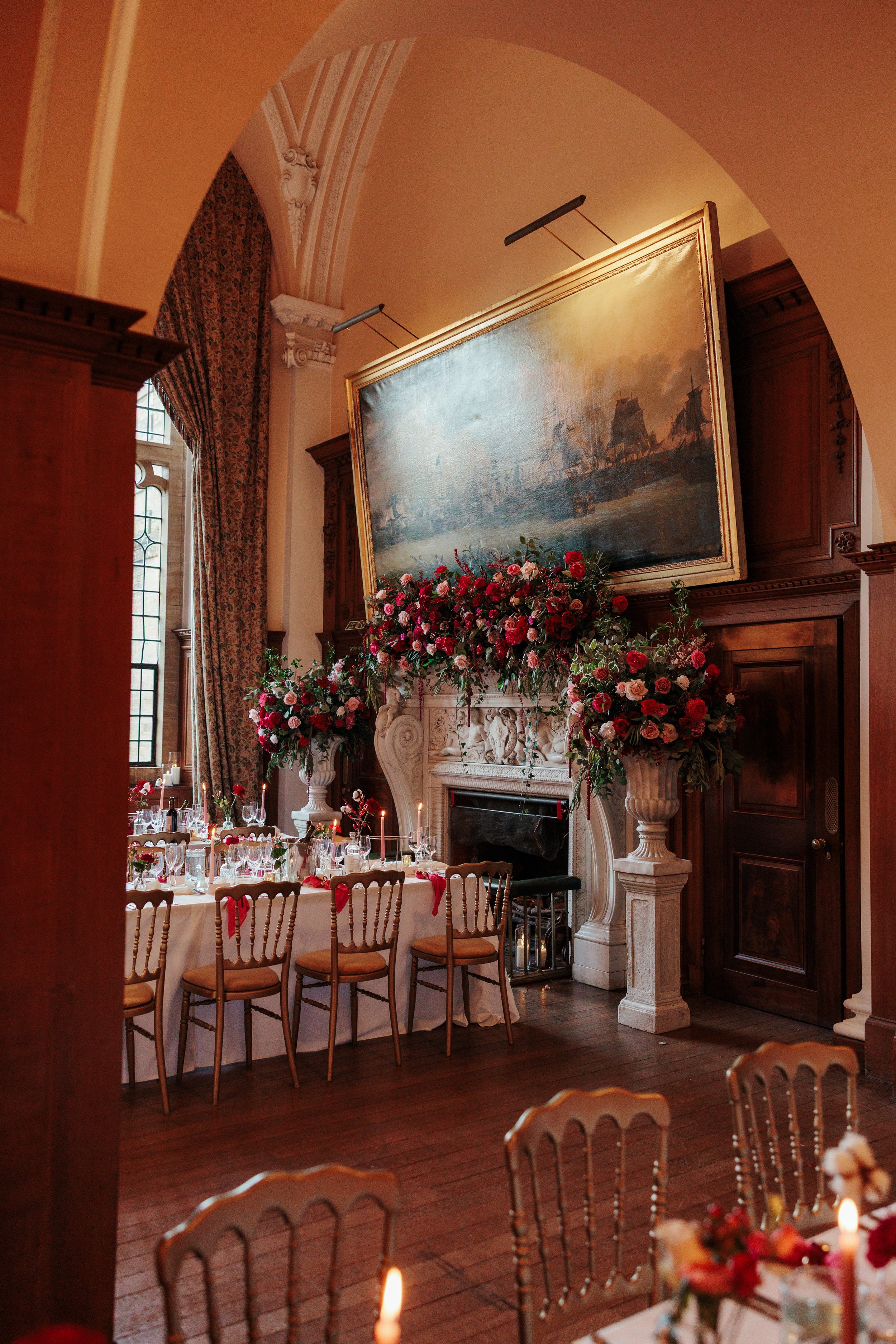An elegant dining room decorated for a celebration with floral arrangements and candles. The room features a large fireplace, a portrait-style painting hung above it, tall windows with patterned curtains, and wooden chairs around a table.