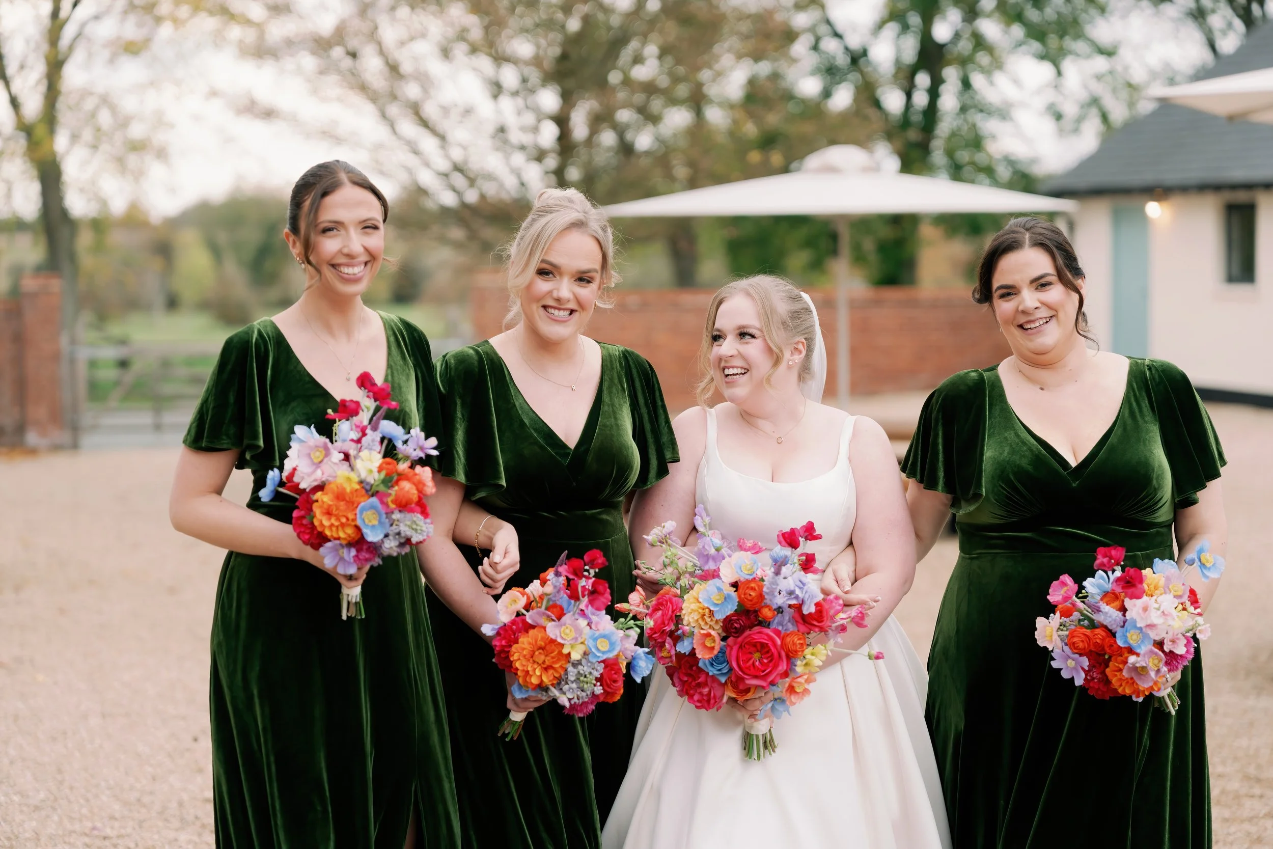 Four women standing outdoors, smiling, dressed in formal attire, holding colorful bouquets of flowers, with a background of trees, a brick wall, and a building with a large umbrella.