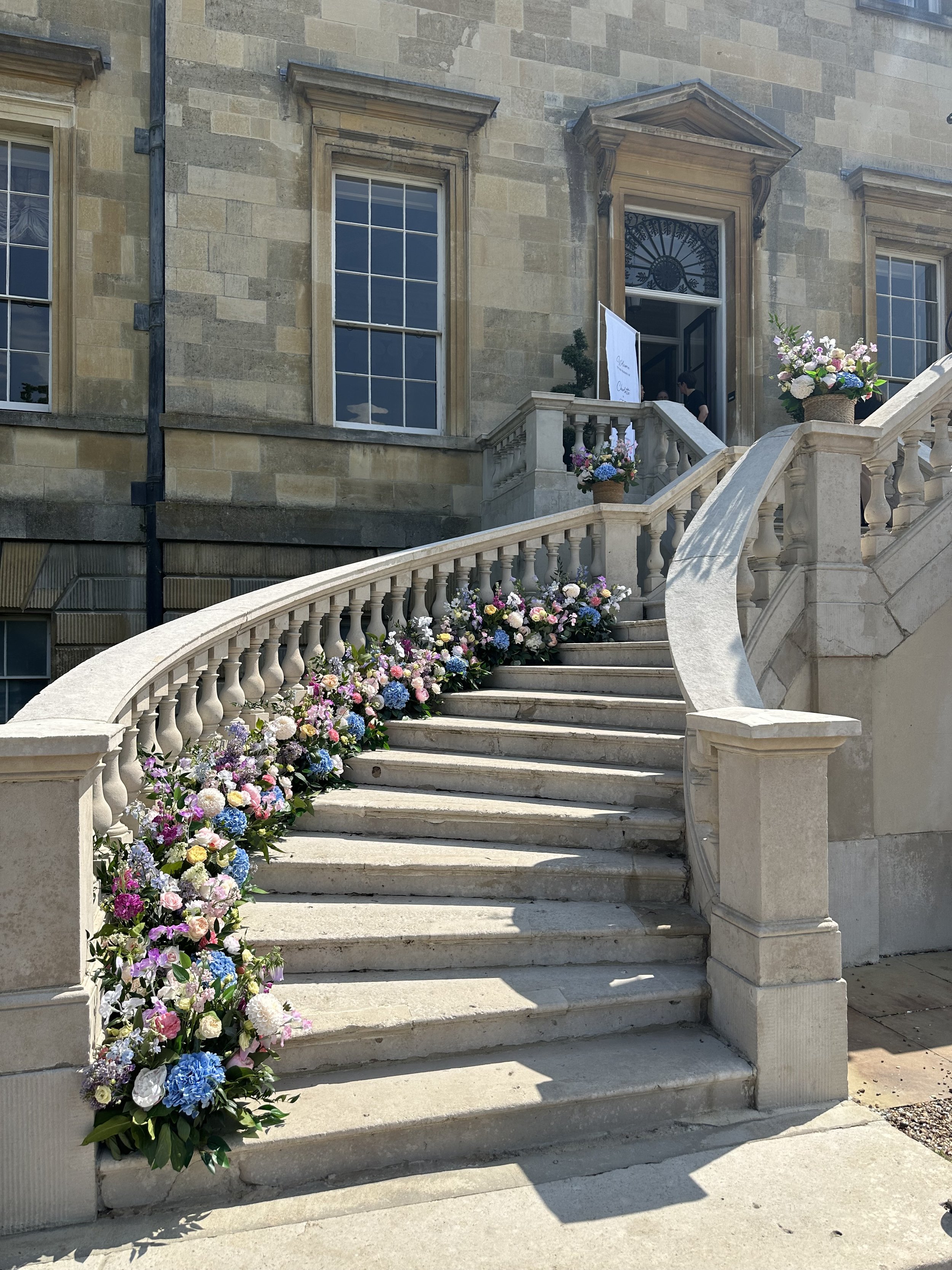 Stone staircase decorated with colorful floral arrangements leading up to the entrance of an elegant, historic building with large windows and classical architectural details.