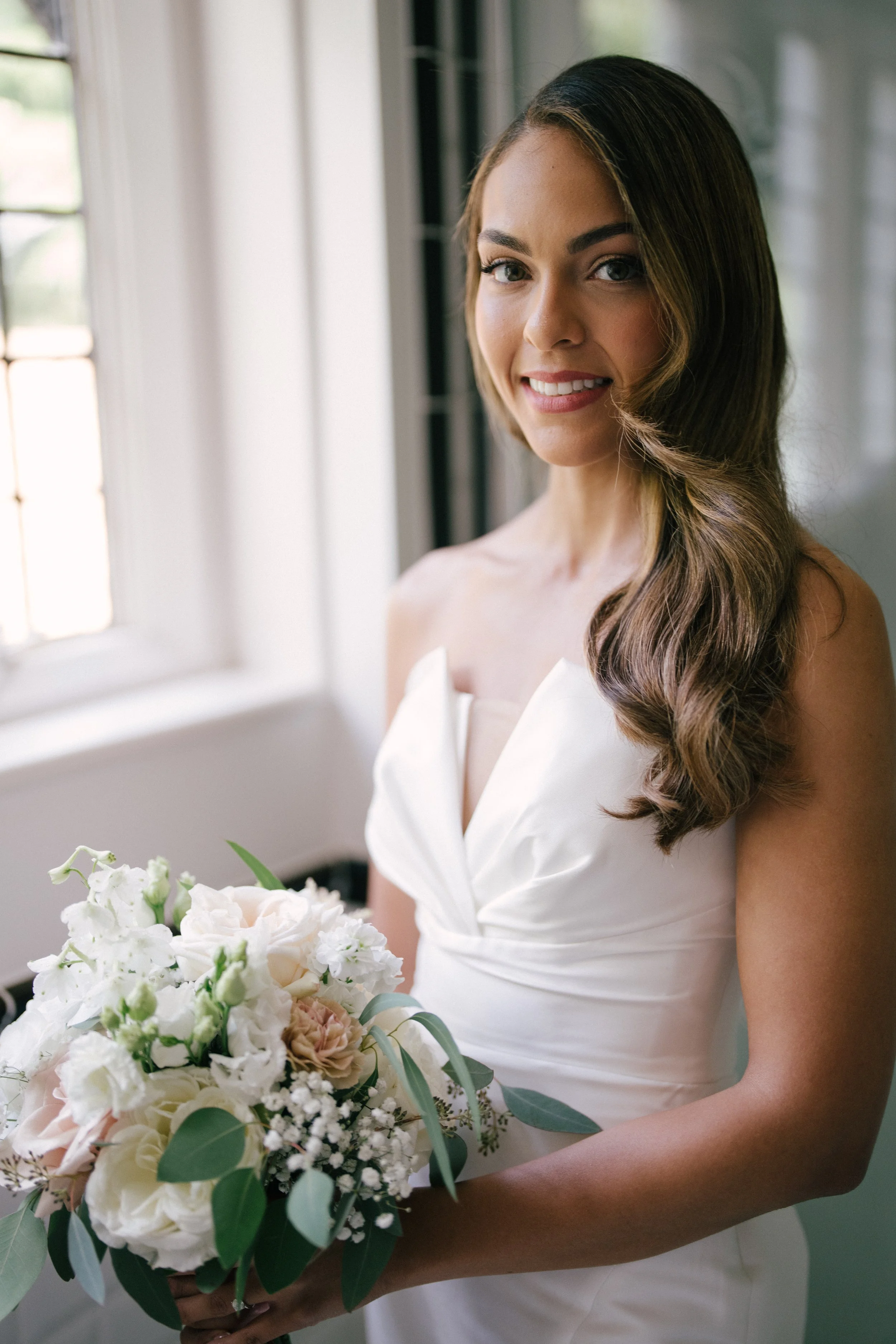 A bride in a white wedding gown holding a bouquet of flowers, standing near a window with sunlight streaming in.