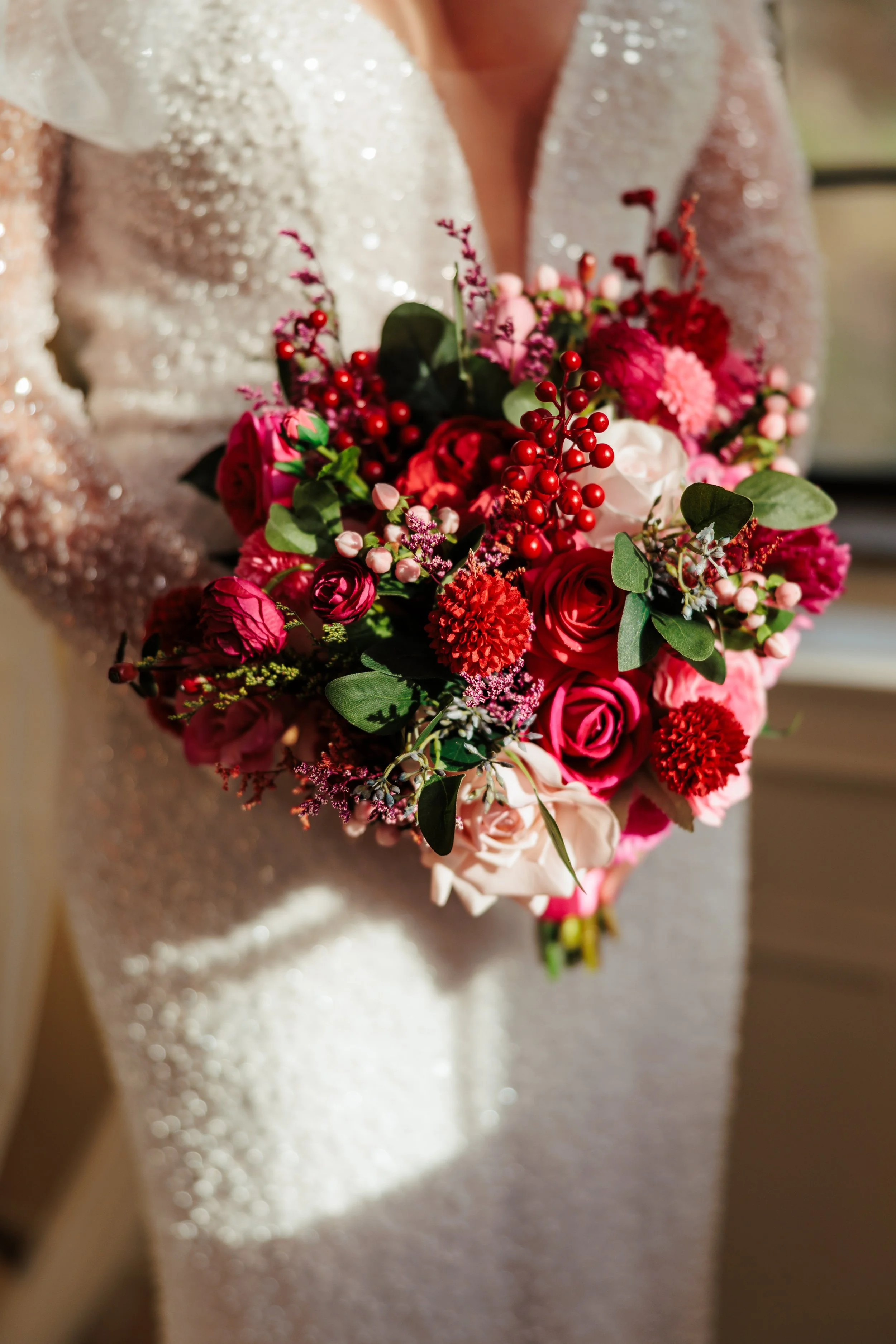 A bride holding a bouquet of pink, red, and white flowers with green leaves in a wedding dress with long sleeves.