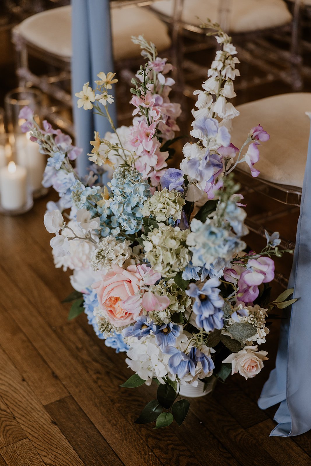 A floral arrangement with pink, blue, white, and purple flowers next to chairs and candles on a wooden floor.