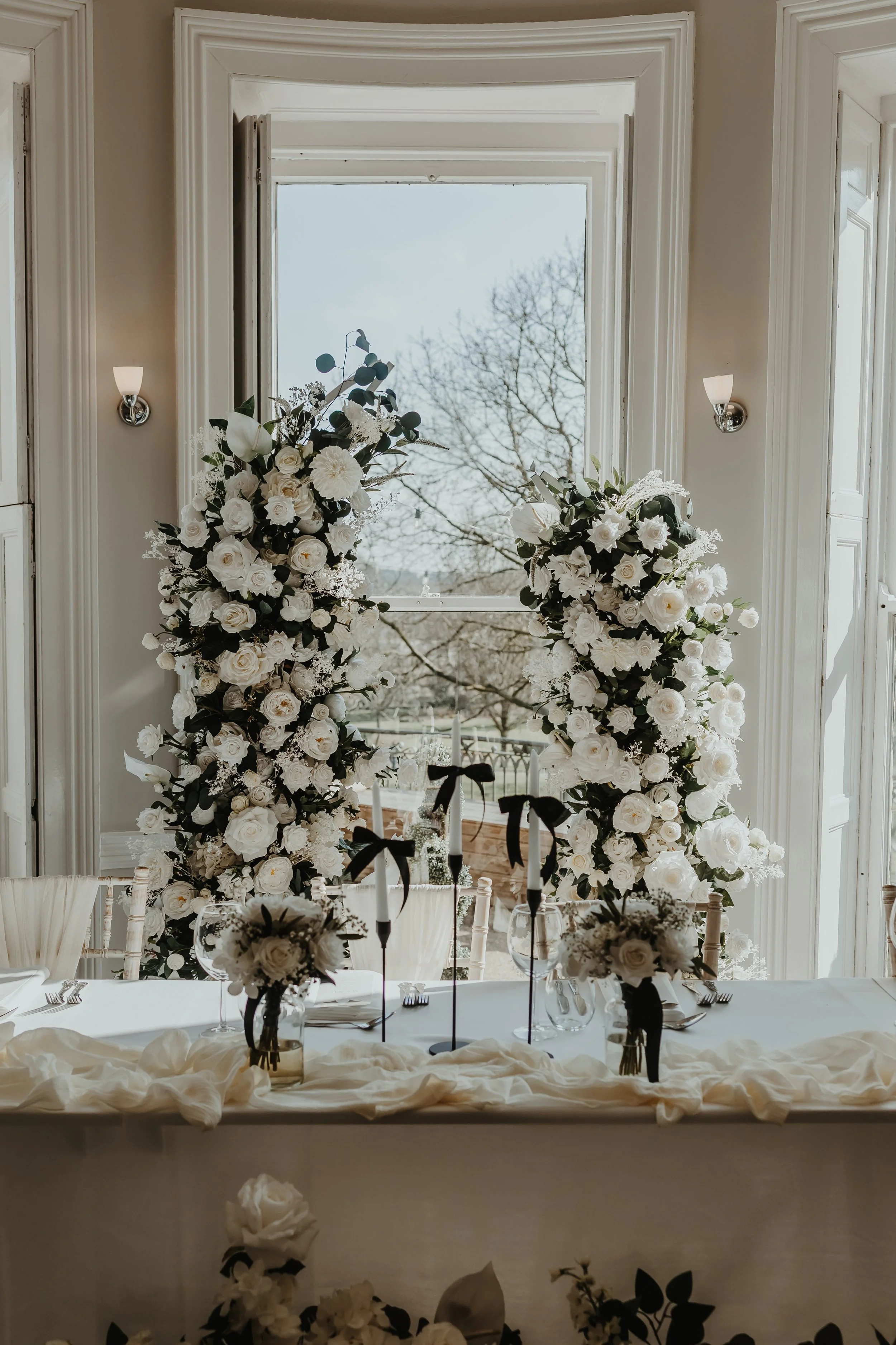 Elegant broken white wedding arch hire using premium faux flowers. Asymmetrical floral installation, styled and installed across Surrey.