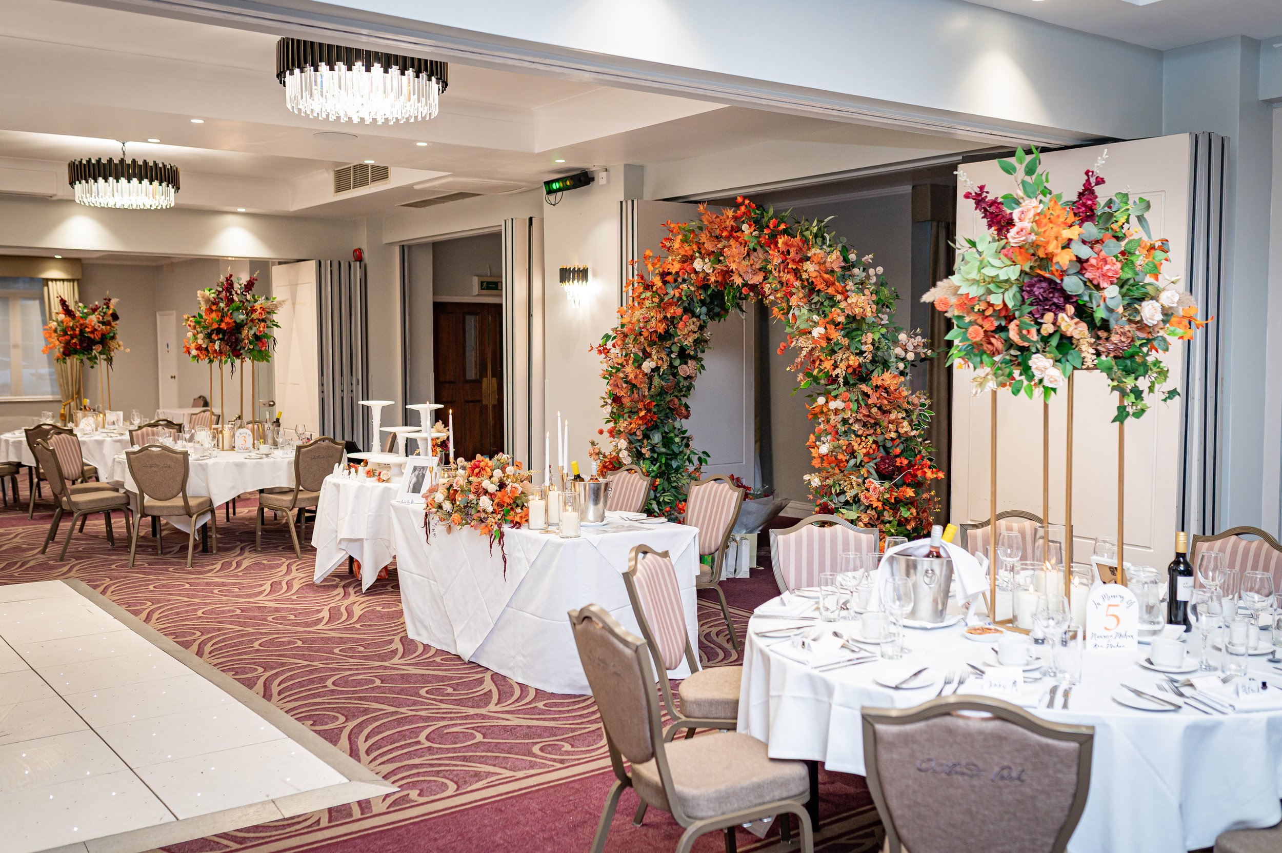 Elegant event space with round tables set for a reception, decorated with floral centerpieces and surrounded by upholstered chairs, featuring a floral archway and modern lighting fixtures.