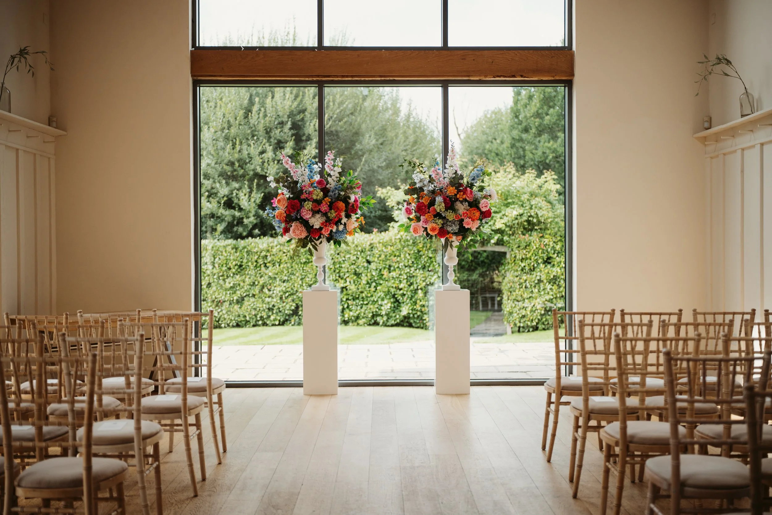 Indoor wedding ceremony setup with two large floral arrangements on white pedestals and rows of wooden chairs facing the front, with a large glass window showing a garden outside.