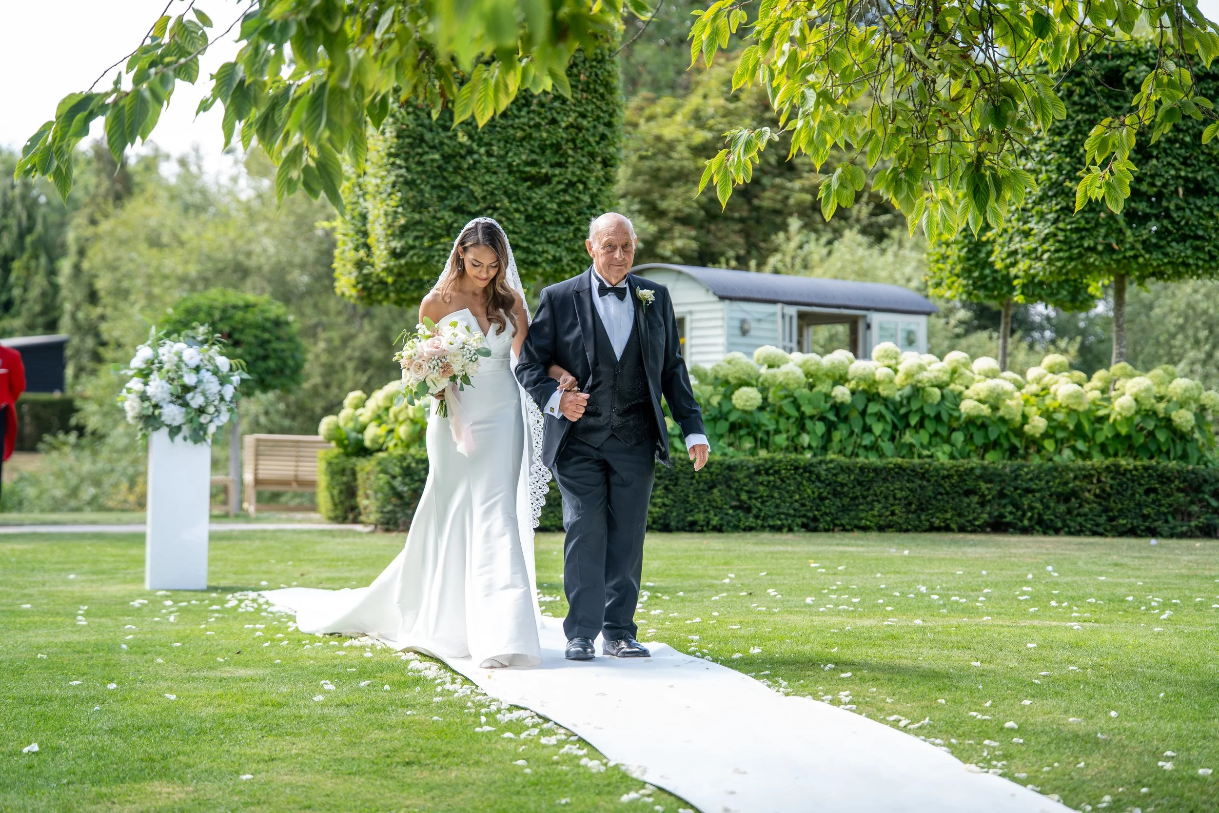 A bride walking arm-in-arm with an elderly man on a white aisle runner during an outdoor wedding ceremony, surrounded by green trees and bushes.