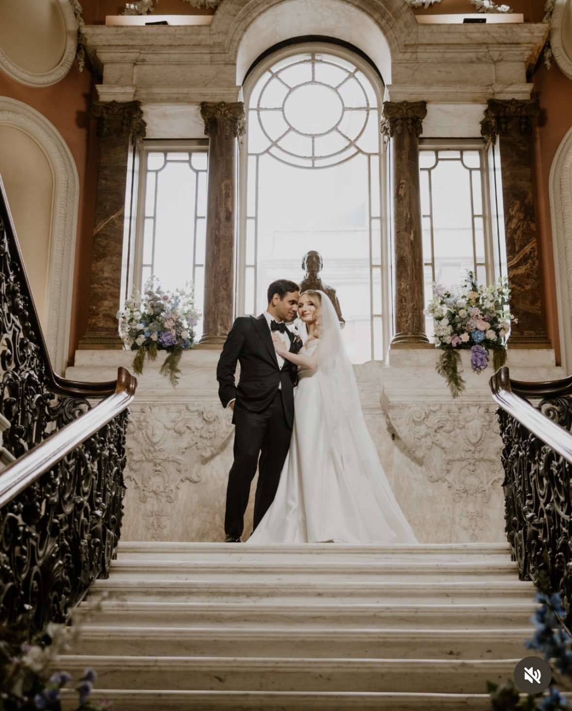 A newlywed couple stands on a grand staircase inside a historic building. The bride wears a white wedding gown and veil, and the groom wears a black tuxedo. Behind them, large windows and a bust sculpture are visible, along with floral arrangements o