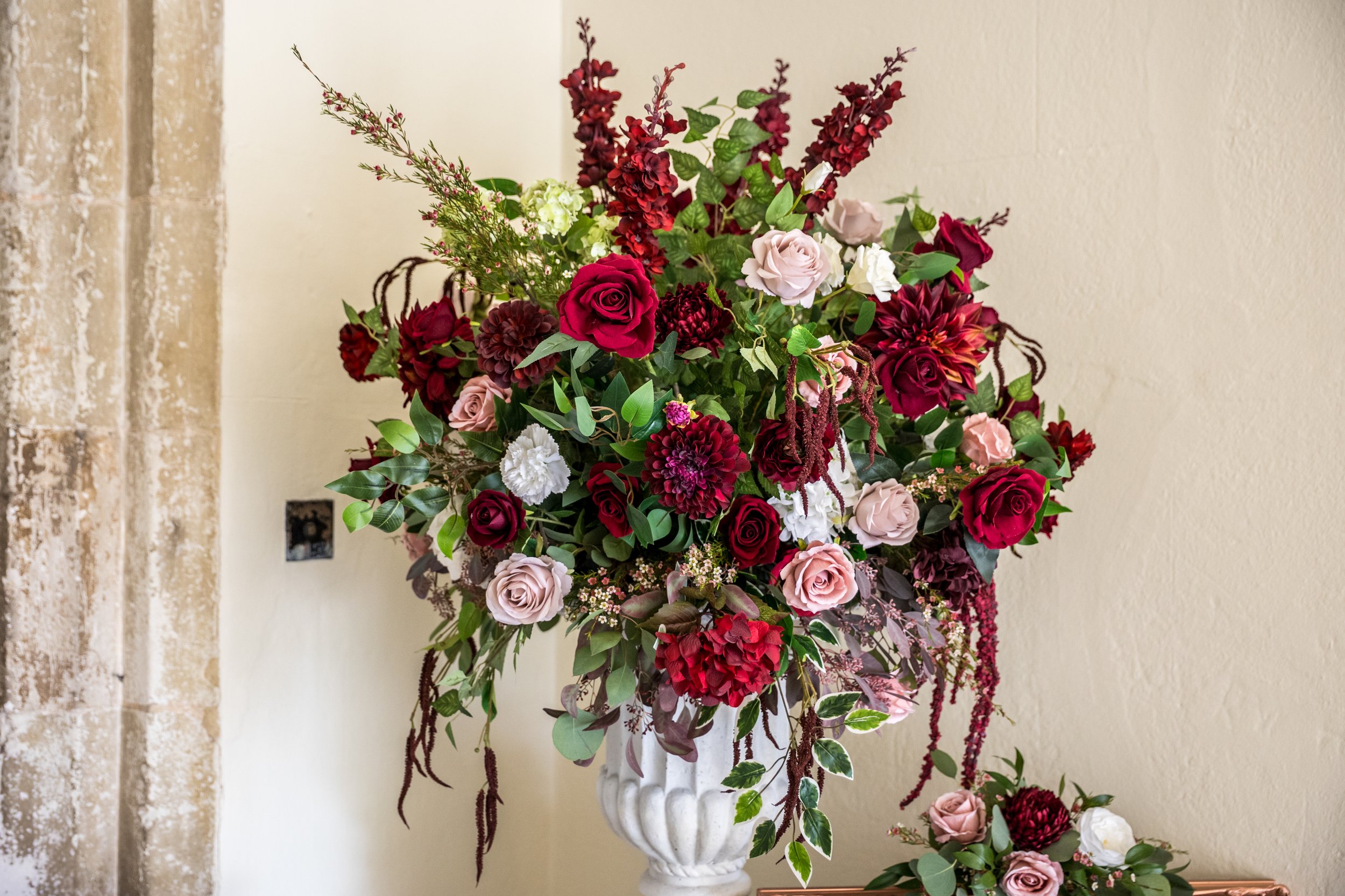 A large floral arrangement in a white vase containing red, pink, burgundy, and white roses, dahlias, and various greenery, placed against a beige wall.