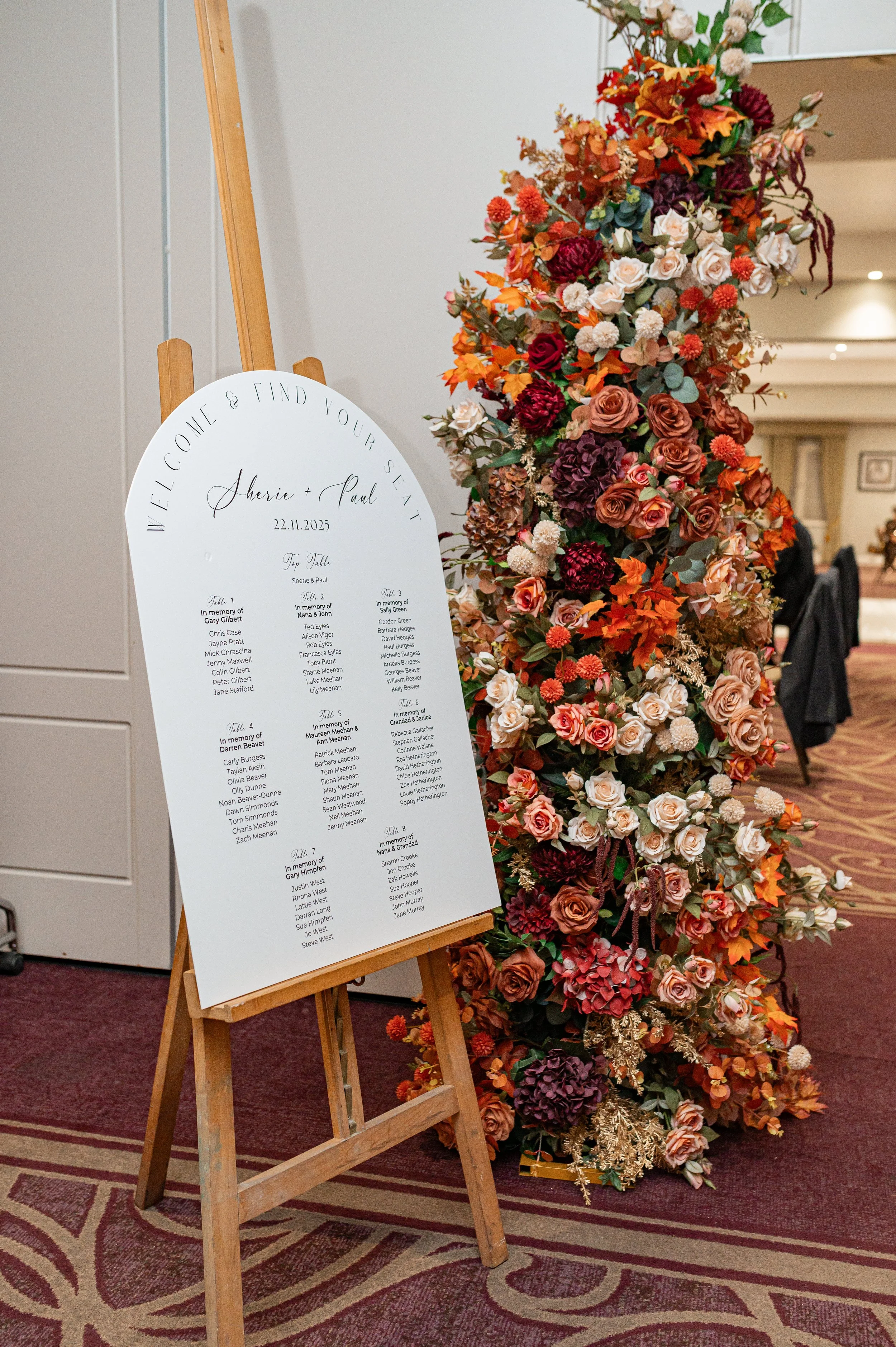 Wedding seating chart and floral display at a reception. The white seating chart board on a wooden easel lists tables and guests' names, with decorative flower arrangement to the right.