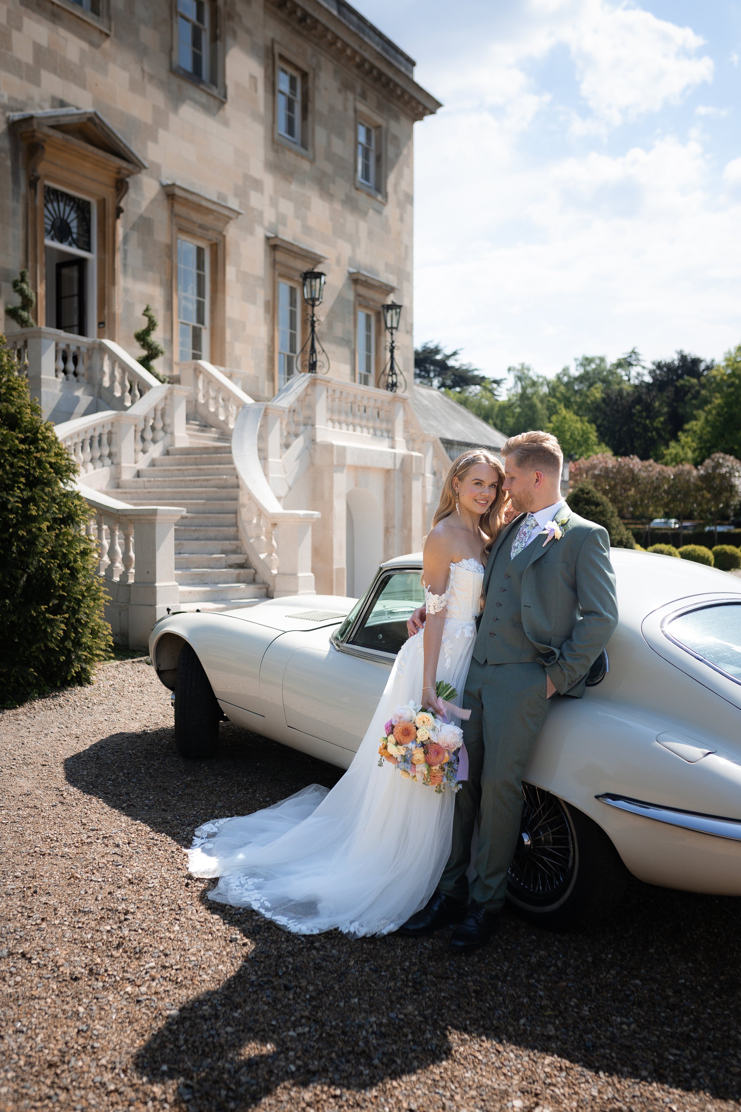 A newlywed couple in wedding attire standing next to a vintage car outside a large estate with a grand staircase and stone facade.