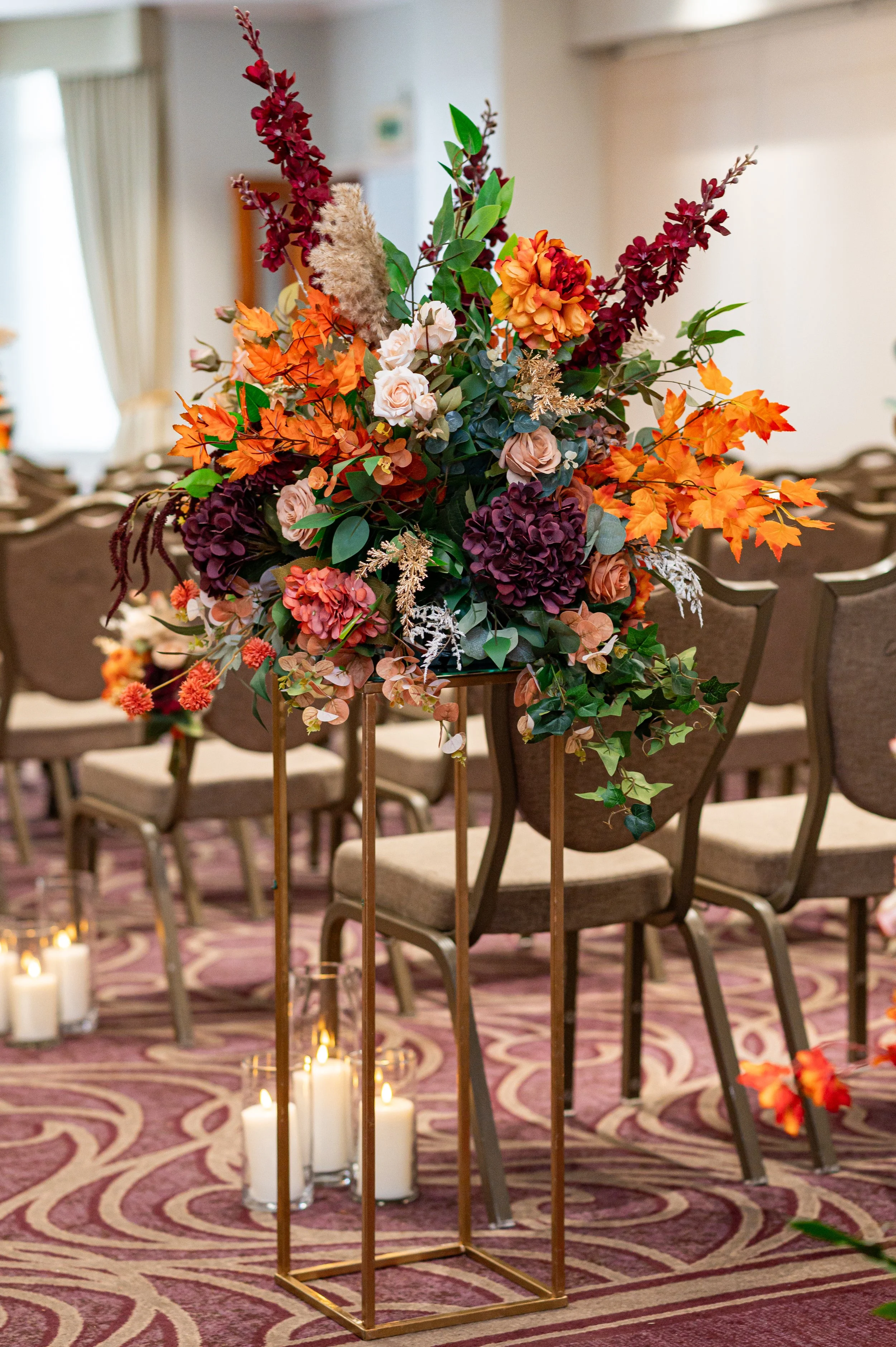 Vibrant fall-themed floral arrangement on a tall gold stand, surrounded by white candles on a patterned carpet.