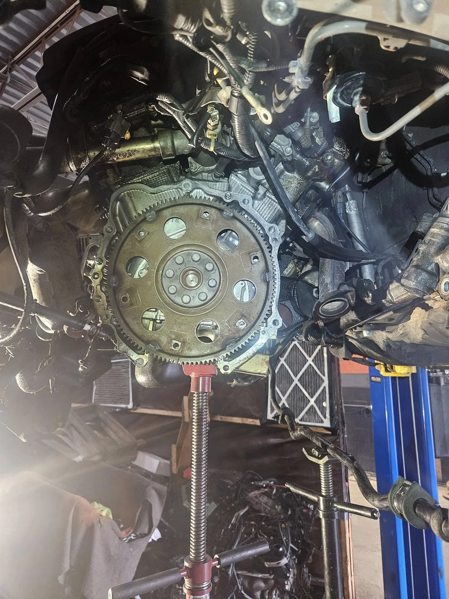 Close-up of a vehicle engine with exposed flywheel and surrounding components, supported on a mechanical stand in a workshop setting.