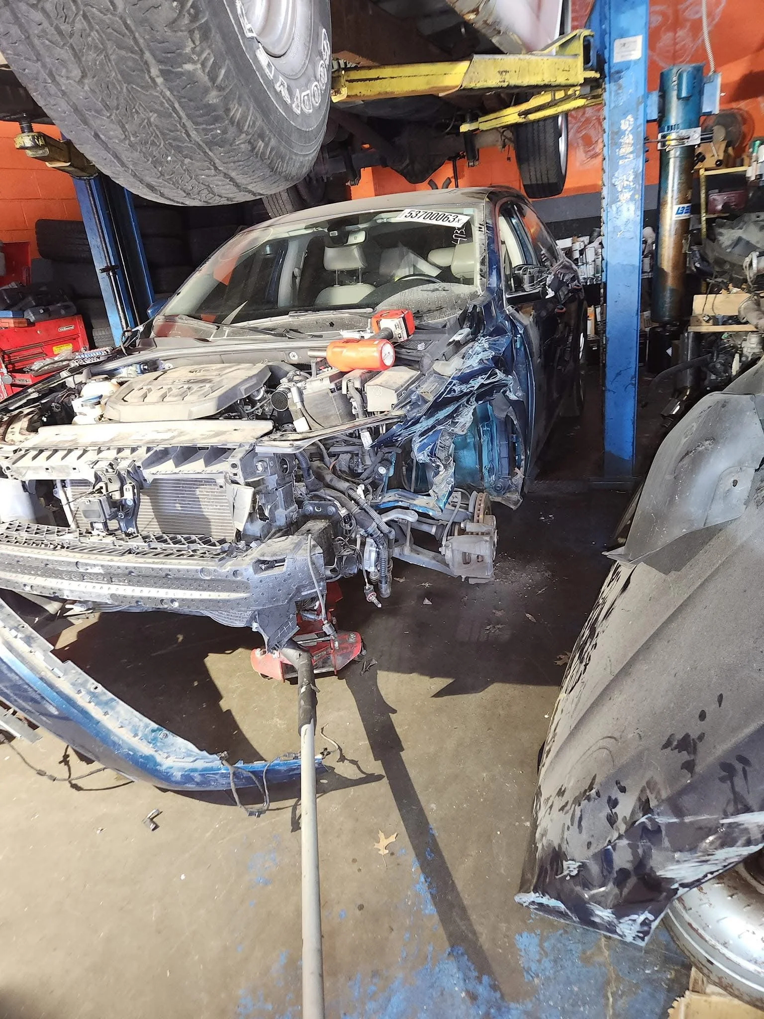 Damaged car in an auto repair shop with parts removed, exposed engine, and nearby car lift.
