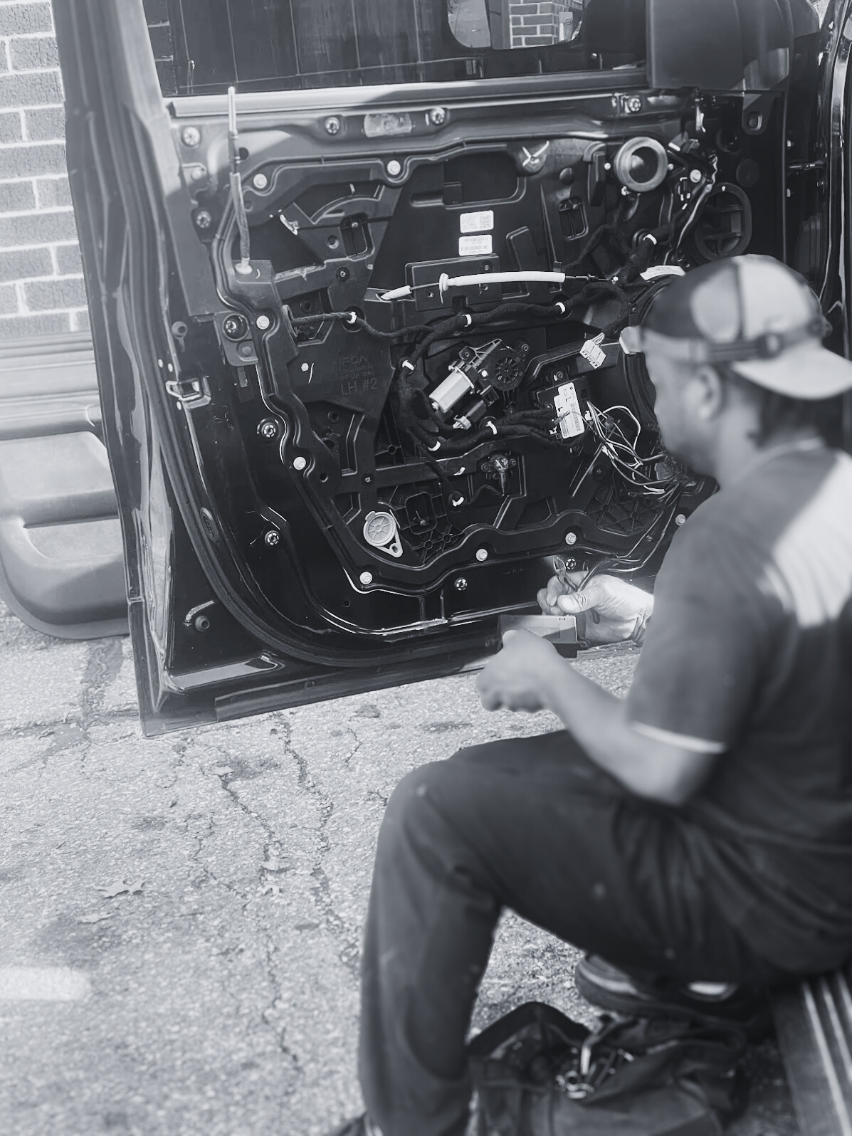 Person working on the inner mechanisms of a car door, wearing a cap and sitting on the ground. The door panel is removed, exposing wires and components.