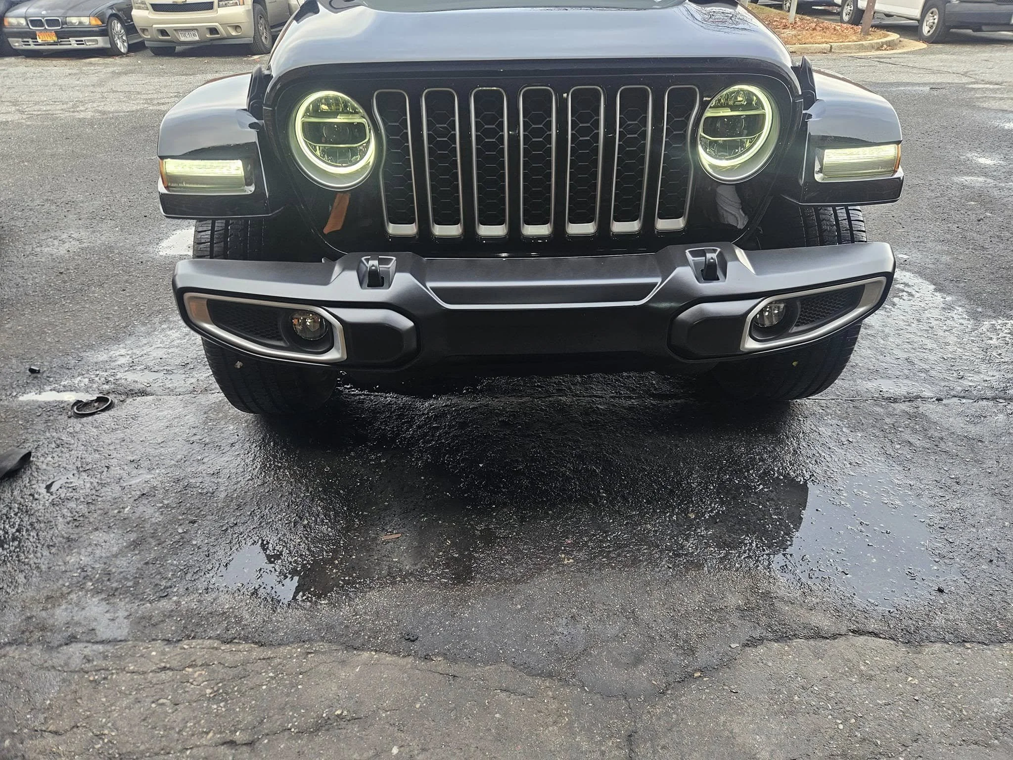 Front view of a black Jeep SUV with round headlights and a seven-slot grille, parked on wet pavement in a parking lot.