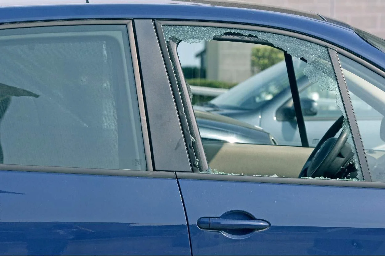 Blue car with a broken front passenger window and shattered glass on the seat, parked in a lot.