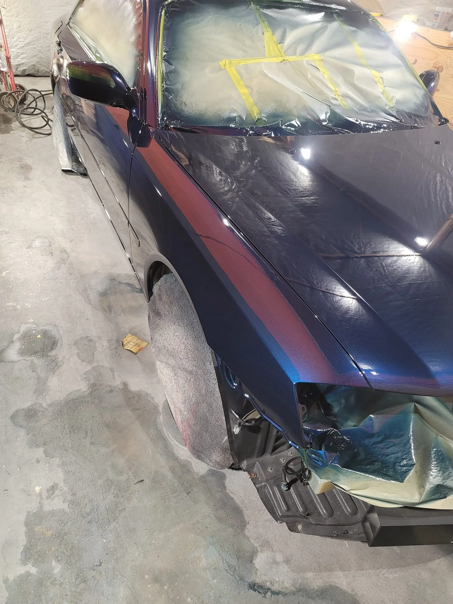 Car being painted with blue color in a workshop. Windshield and wheels are covered for protection during painting process.