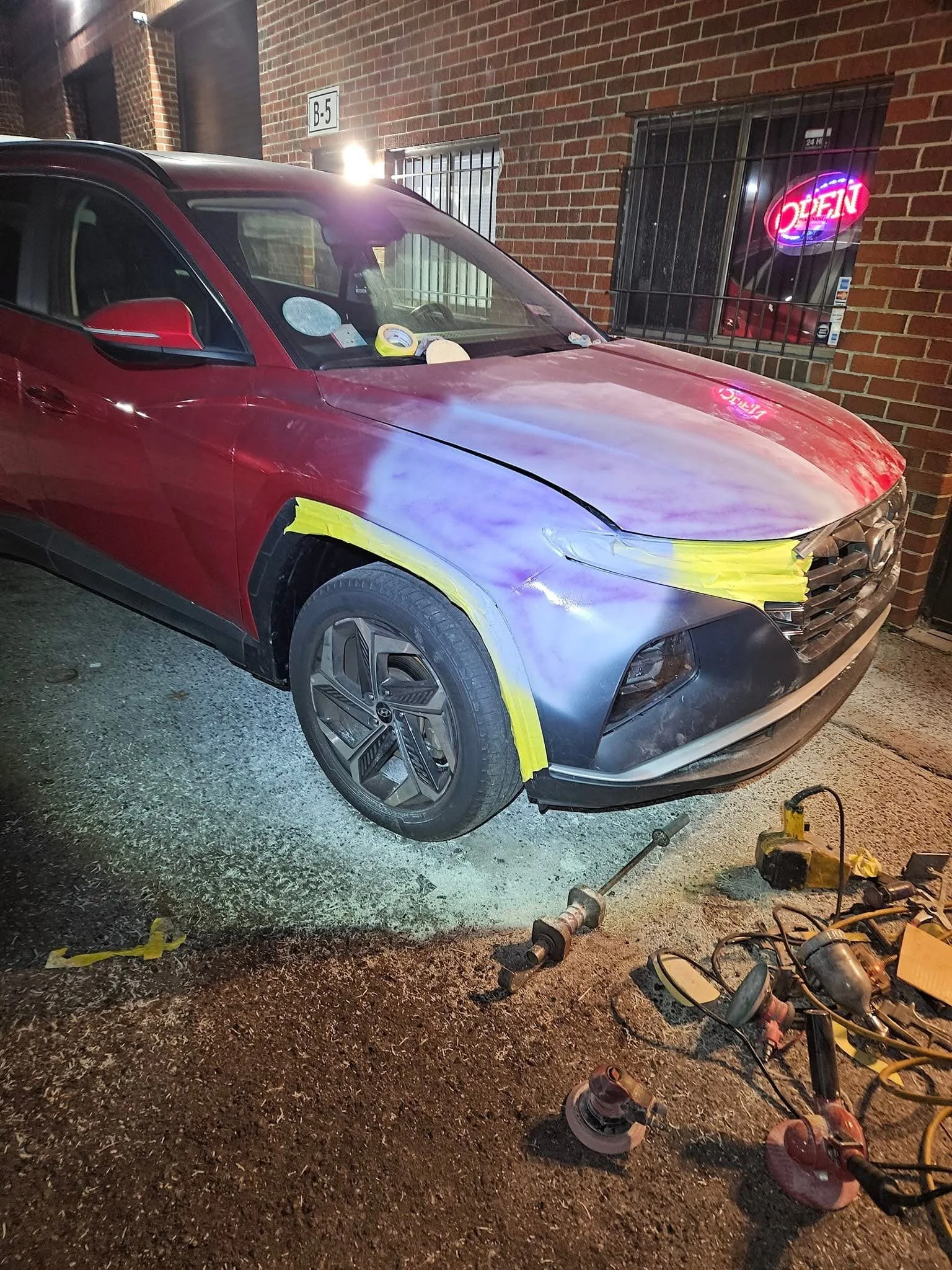 A red SUV with a partially sanded front fender undergoing bodywork repair. The fender is taped off with yellow tape and tools are scattered on the ground nearby. A brick wall with a neon 'Open' sign is in the background.