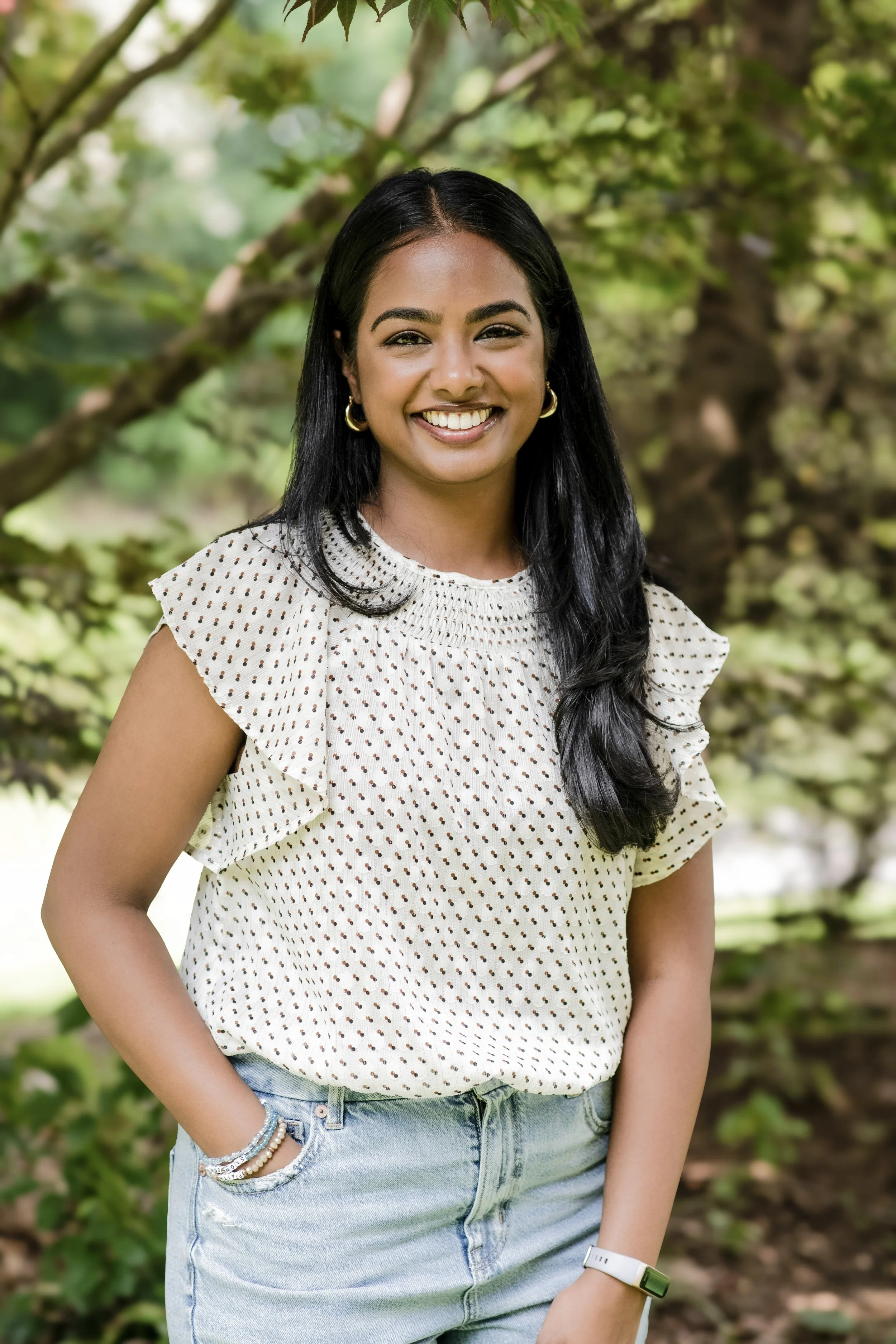 A young woman with long black hair, wearing a white polka dot blouse and light blue jeans, smiling outdoors in a park with green trees in the background.
