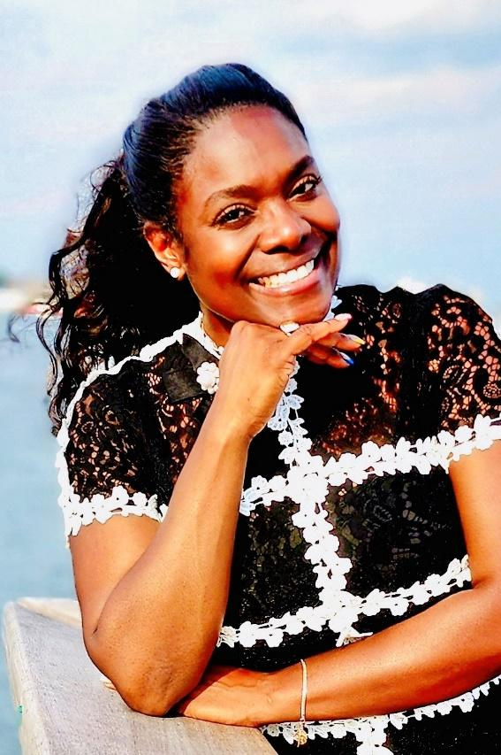 A woman with dark hair in a ponytail smiling, resting her chin on her hand, wearing a black lace top with white floral accents, outdoors near water with a cloudy sky in the background.