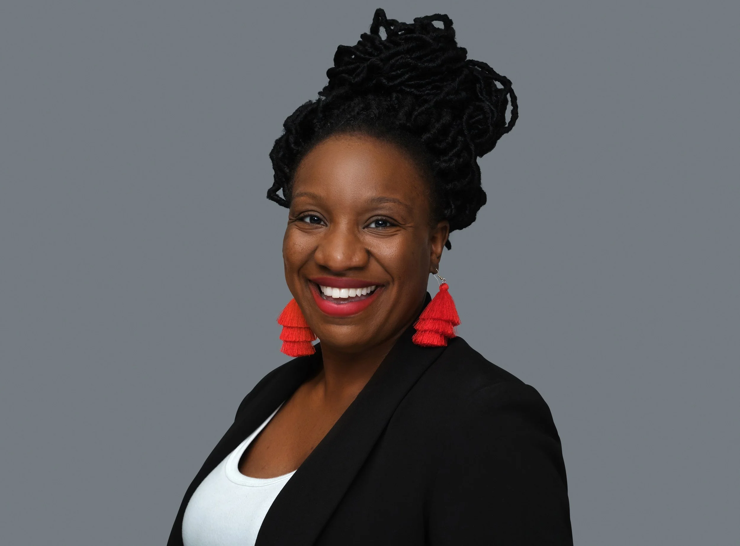 A woman with dark skin, wearing a black blazer, white top, and red tassel earrings, smiling against a gray background.