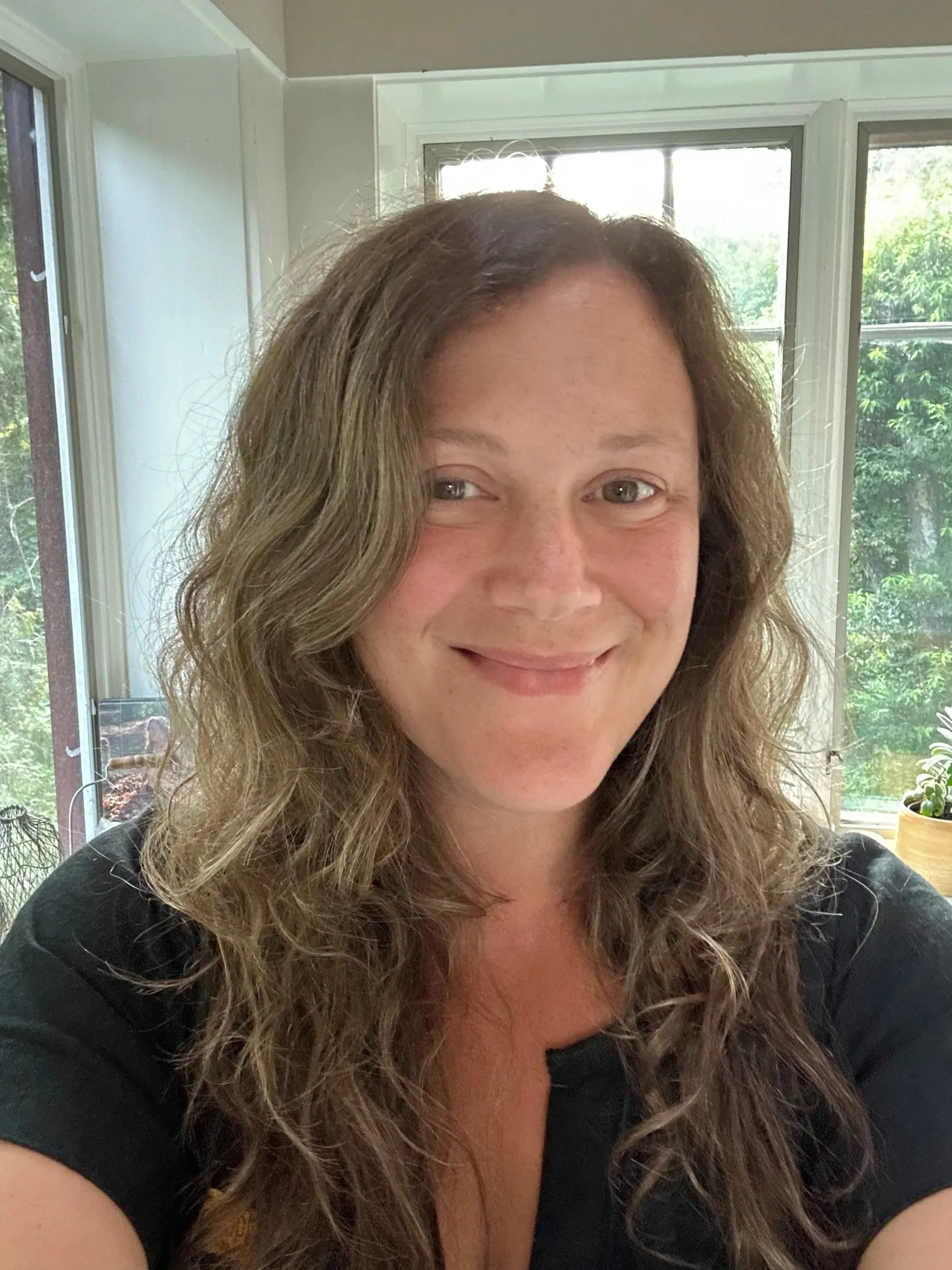 A woman with long, curly brown hair smiling inside a sunlit room with large windows showing green trees outside.