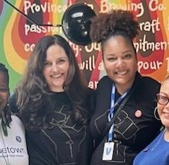 Three women smiling, standing together at an event with a colorful background and microphones.