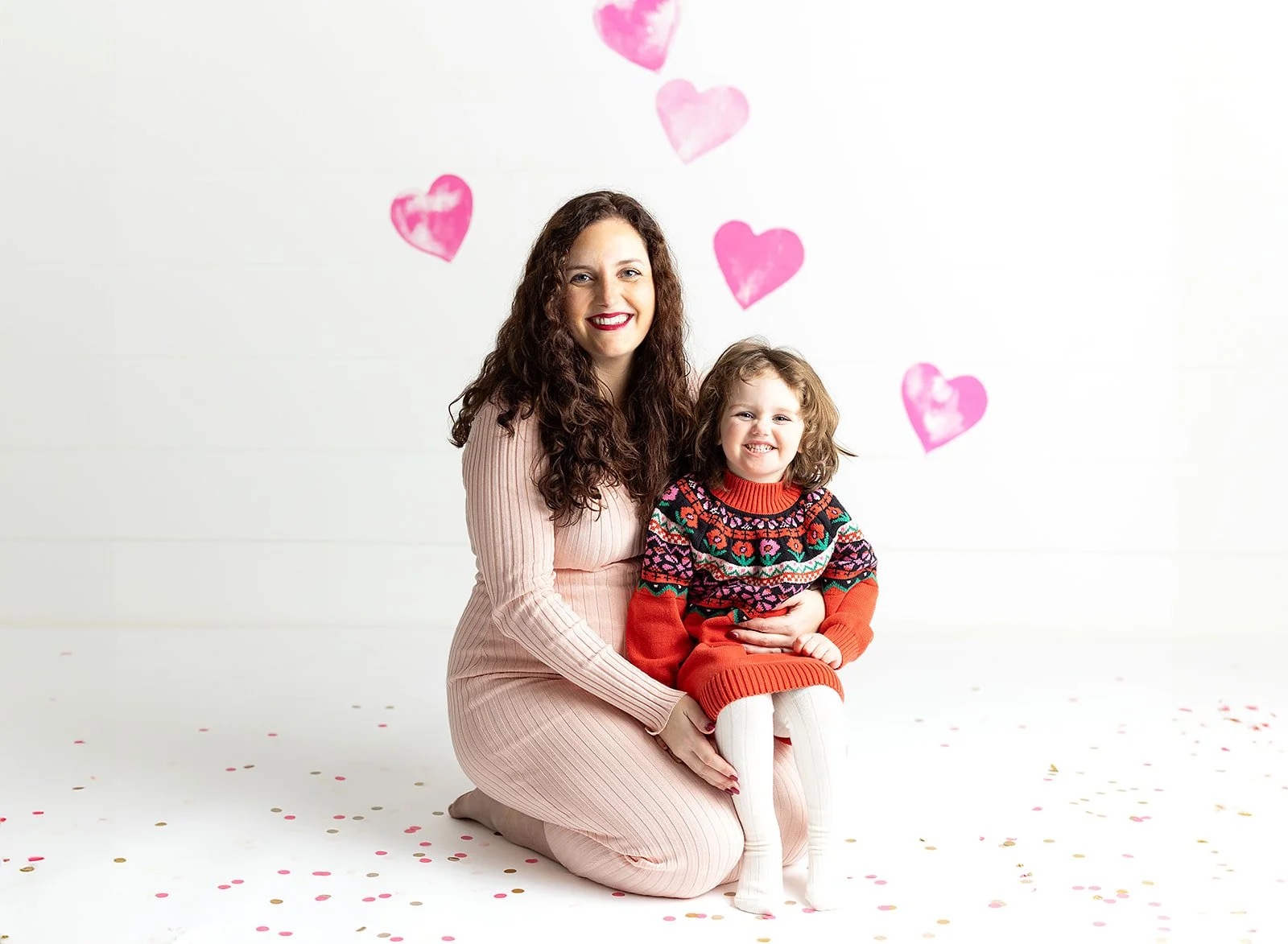 A smiling woman with long curly brown hair kneeling beside a young girl with curly light brown hair. They are in front of a white background decorated with pink heart-shaped balloons and scattered confetti.