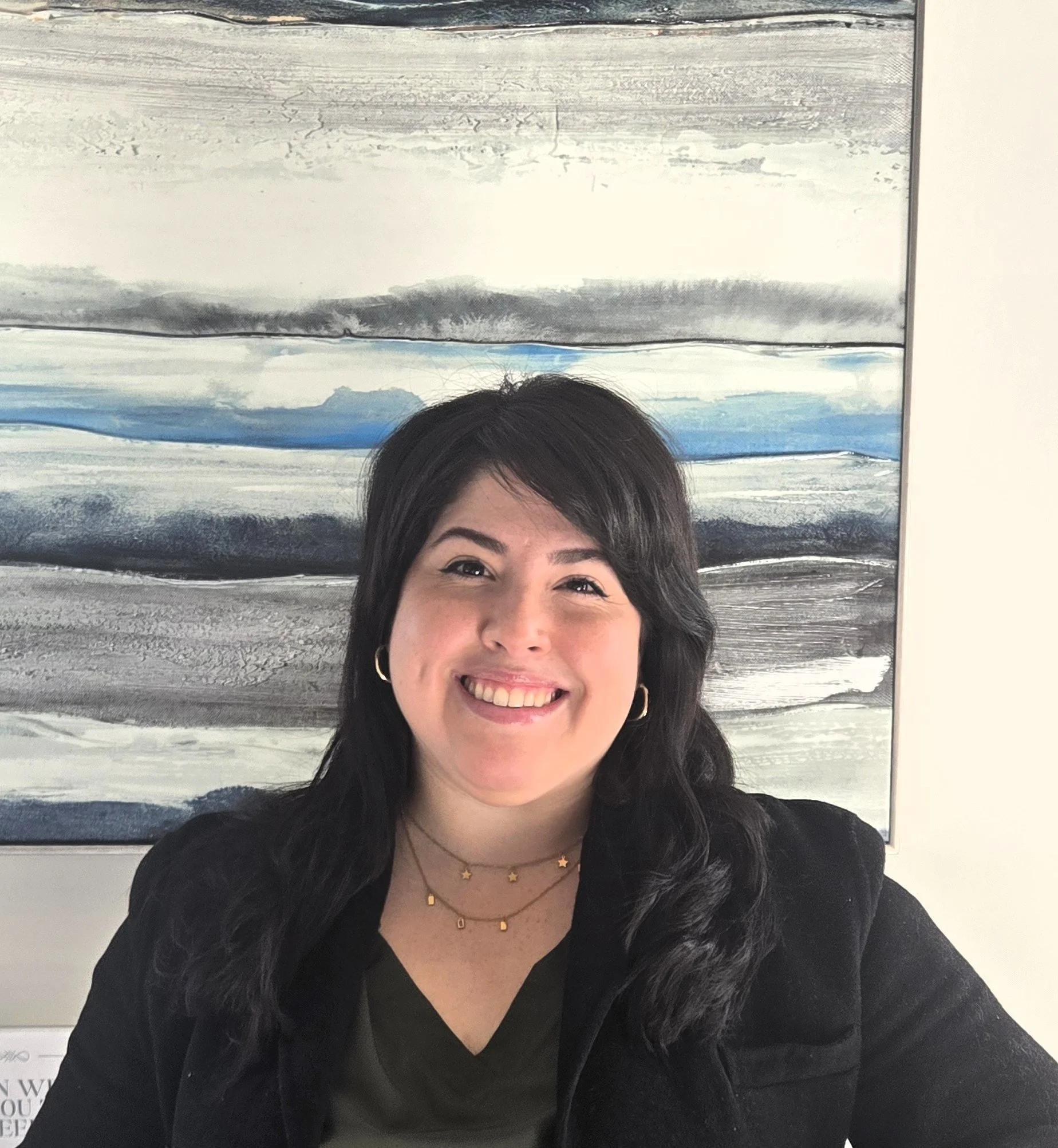 Smiling woman with dark hair, wearing gold hoop earrings, layered necklaces, a black top, and a black jacket, standing in front of an abstract painting with shades of gray, white, and blue.