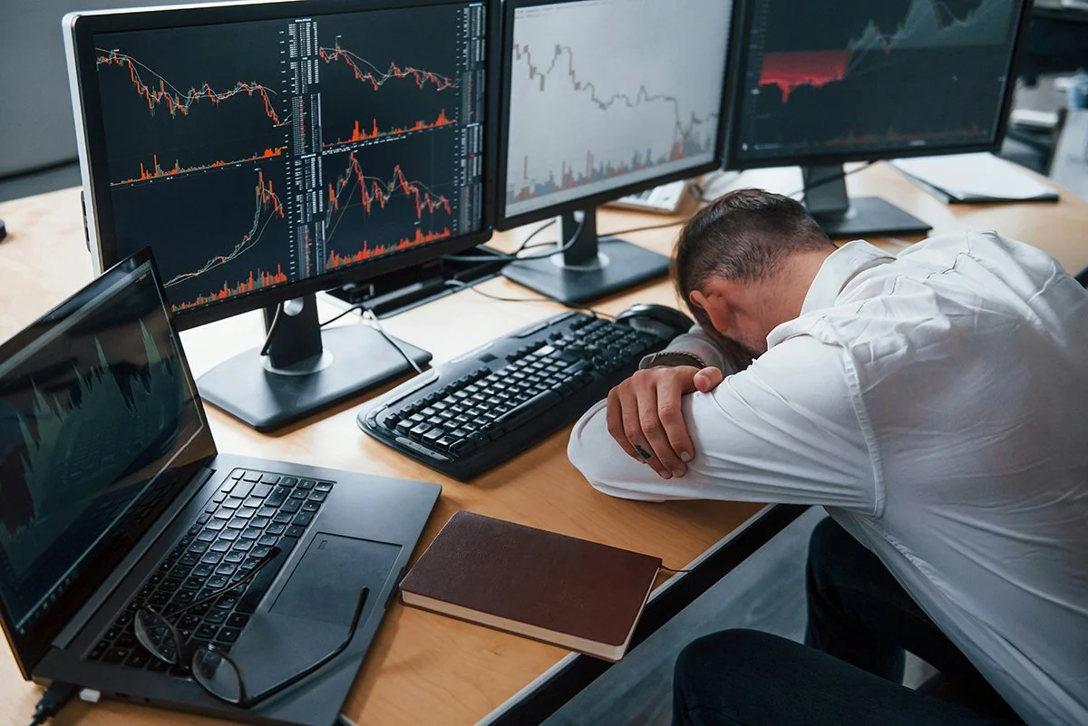 Man with head down on a desk surrounded by multiple monitors displaying stock market charts and data, a laptop, a keyboard, a closed notebook, and glasses.