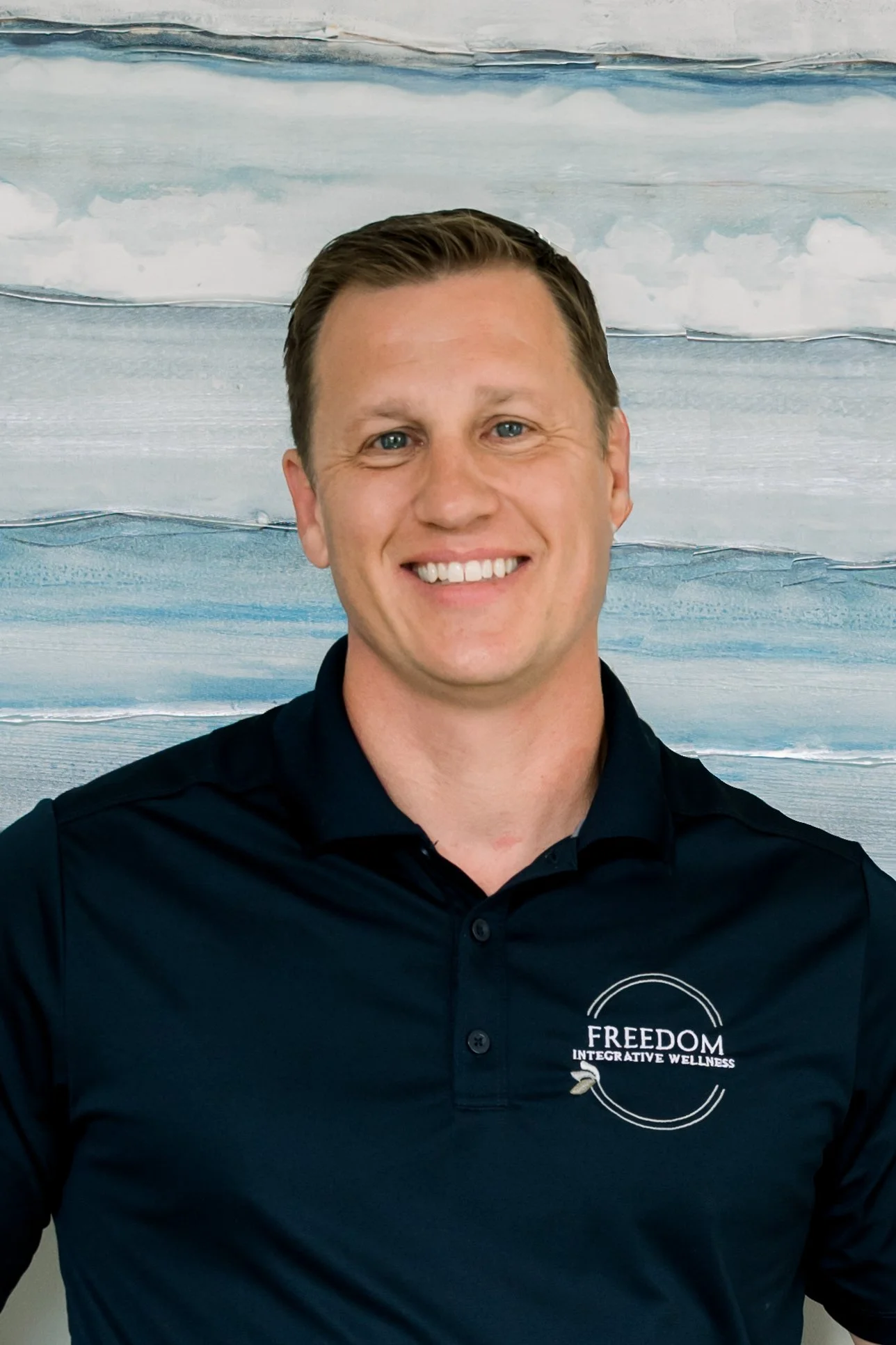 Smiling man in navy polo shirt with 'FREEDOM INTEGRATIVE WELLNESS' logo, standing in front of a sea-themed painting with blue and white waves.
