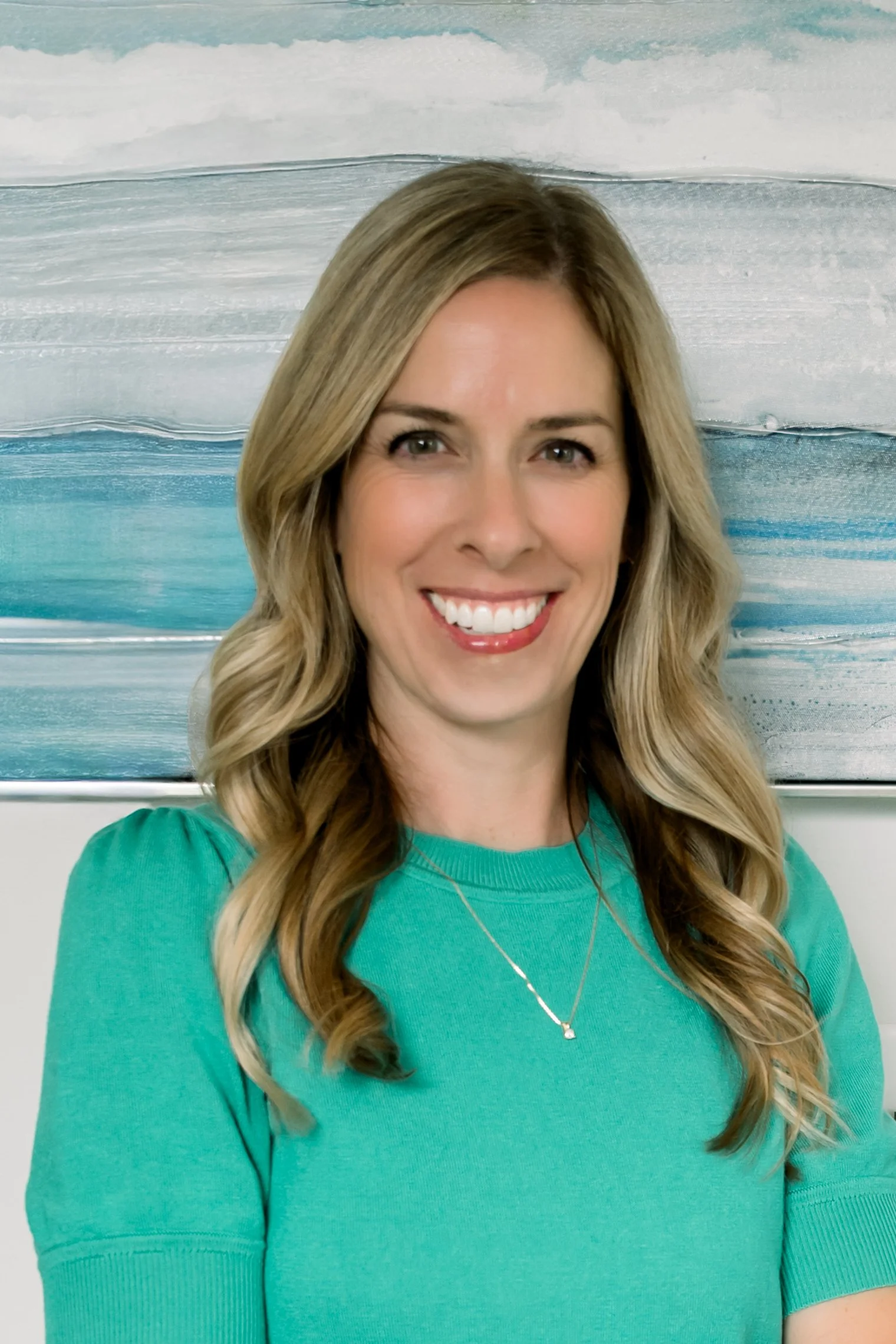 A woman with long wavy blonde hair, wearing a turquoise shirt and a delicate necklace, smiling in front of an abstract painting with shades of blue and white.