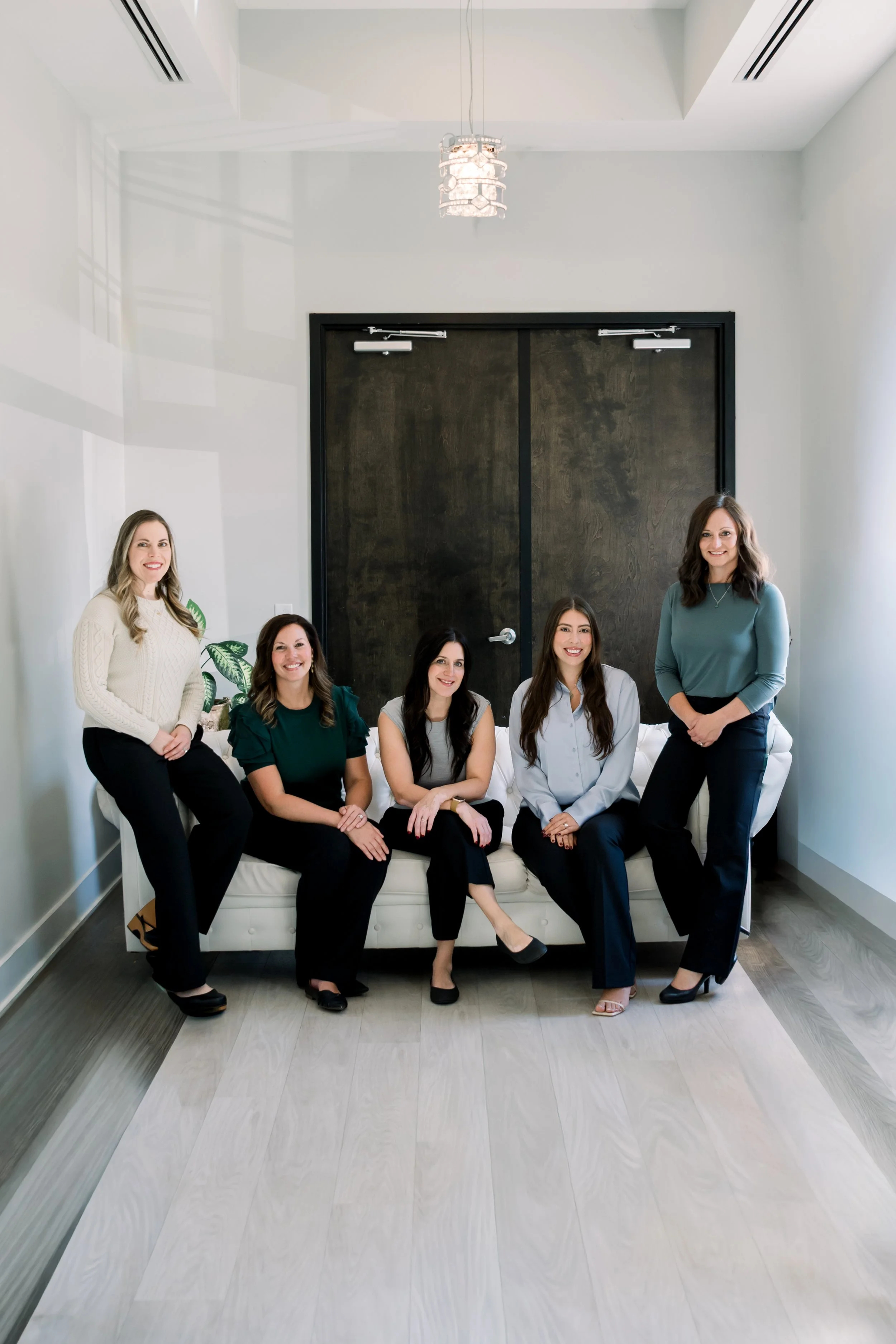 Five women dressed in business casual attire sitting and standing on a white sofa in a modern room with white walls and dark wood flooring.