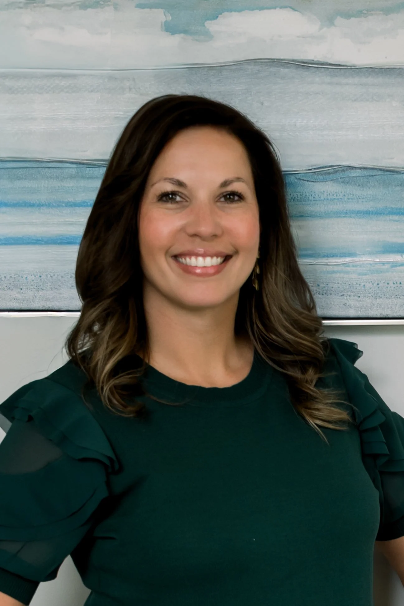 A woman with shoulder-length wavy brown hair, smiling, wearing a dark green top with ruffled sleeves, standing in front of a abstract blue and gray painting.
