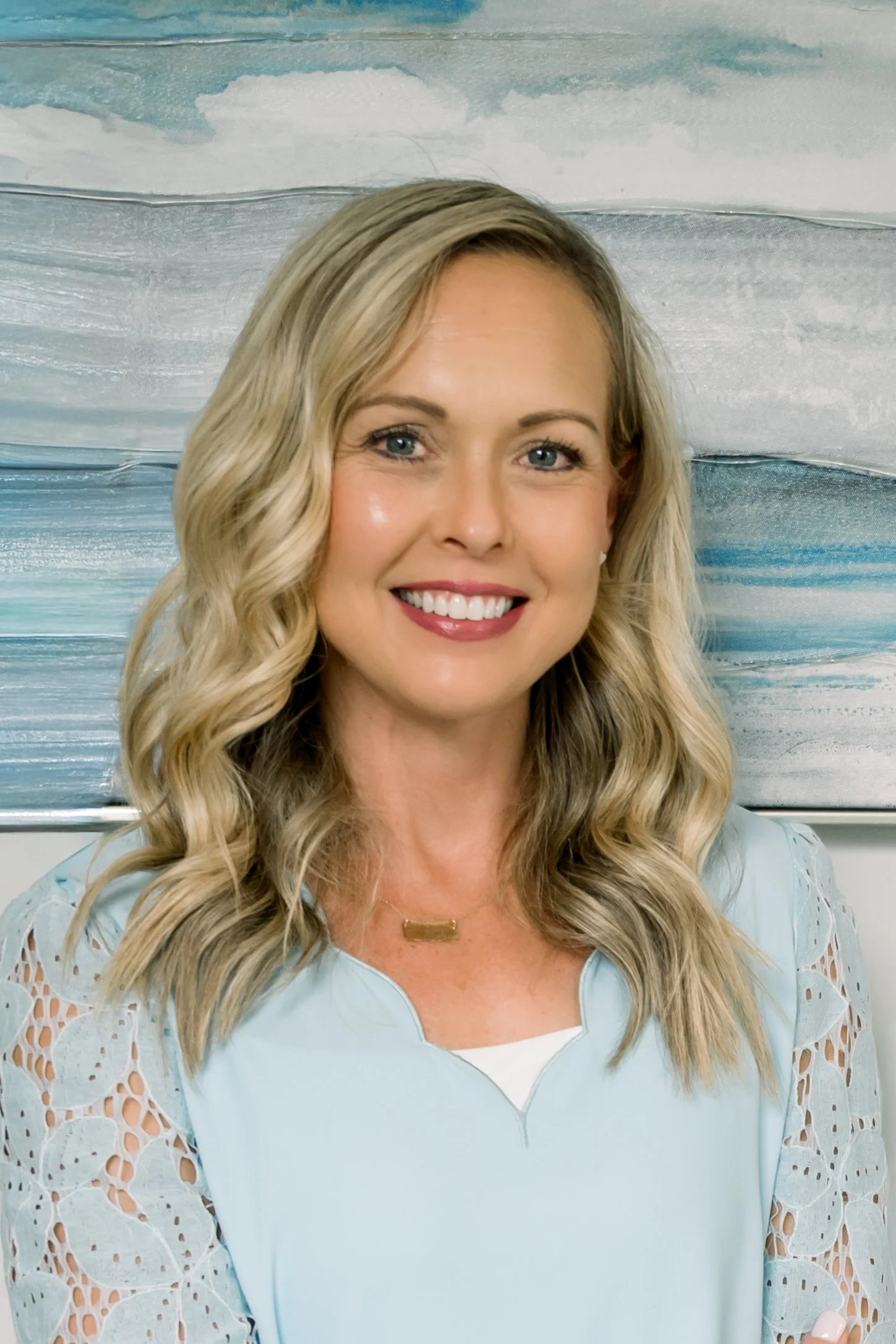 Portrait of a smiling woman with wavy blonde hair, wearing a light blue top with lace sleeves, standing in front of an abstract blue and white background.