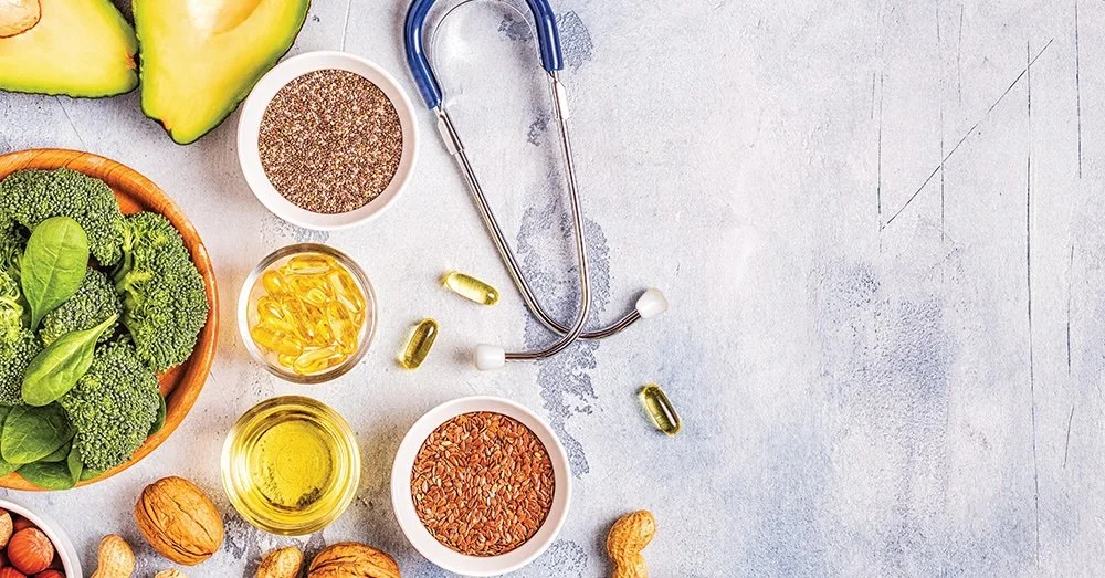 Healthy foods and supplements on a light surface, including avocado, broccoli, walnuts, flax seeds, fish oil capsules, and a stethoscope.