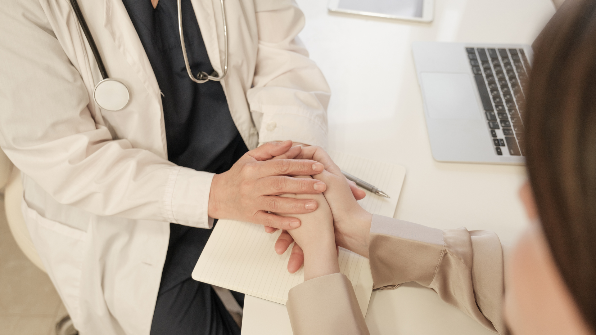 Doctor holding patient's hand during consultation, with notebook, pen, and laptop on table.