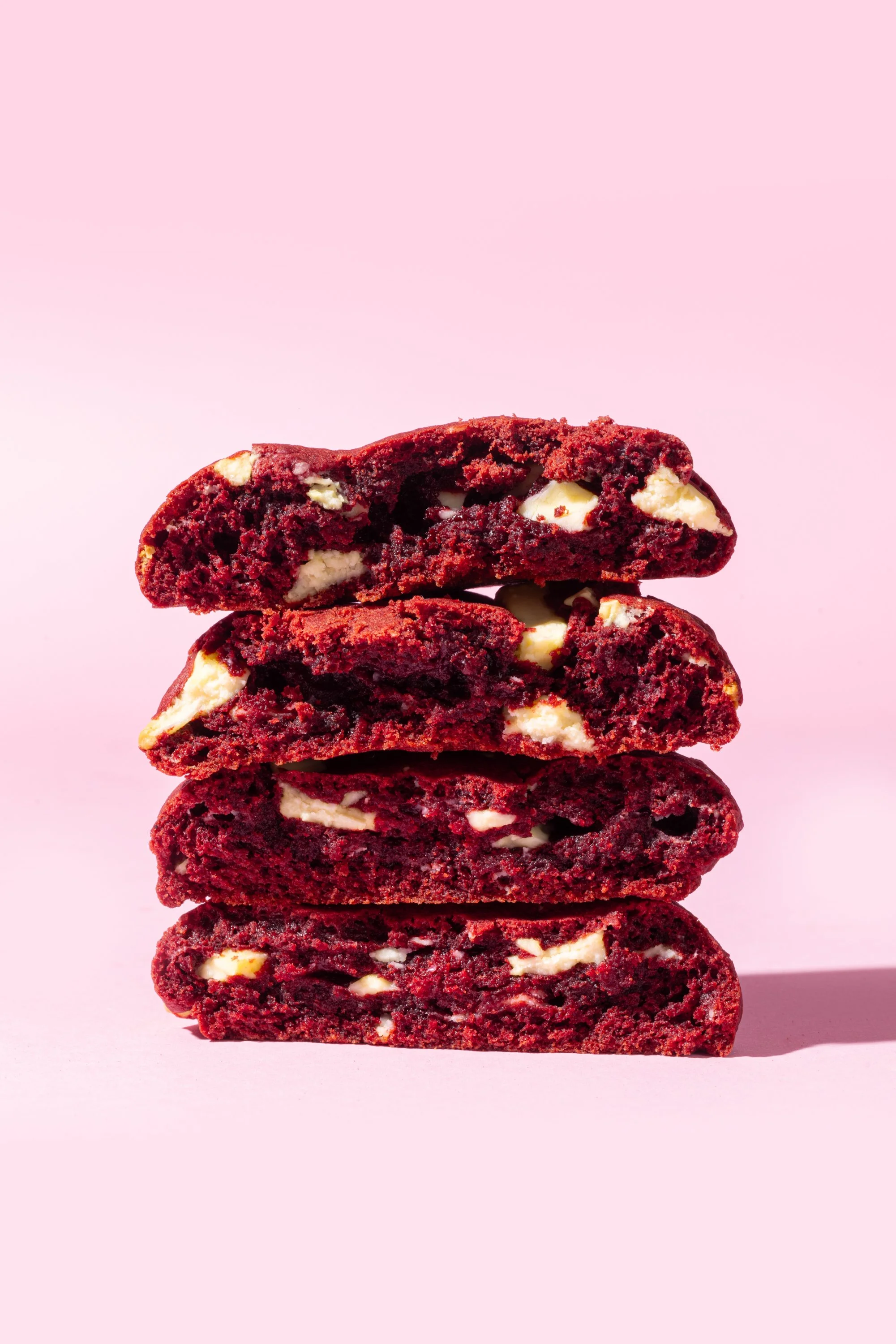 Stack of three red velvet cookies with white chocolate chips on a pink background.