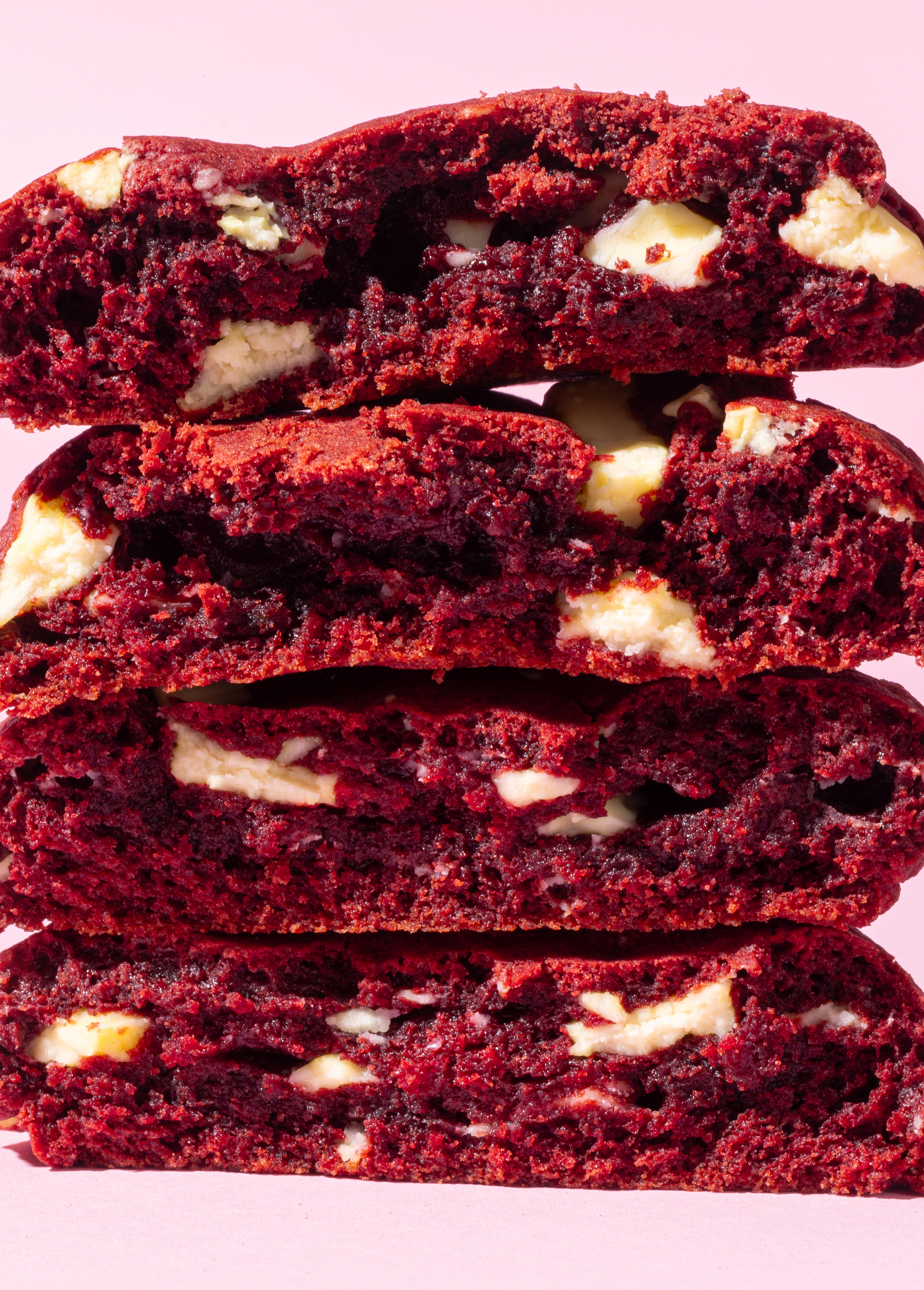 Stack of four red velvet cookies with white chocolate chunks on a pink background.