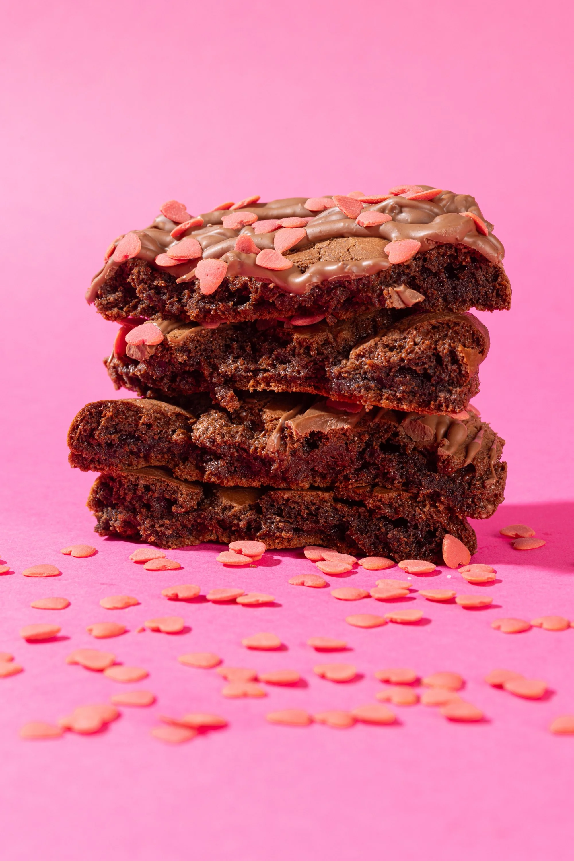 Stack of three chocolate cookies with pink heart-shaped sprinkles on a pink background, with more sprinkles scattered around.