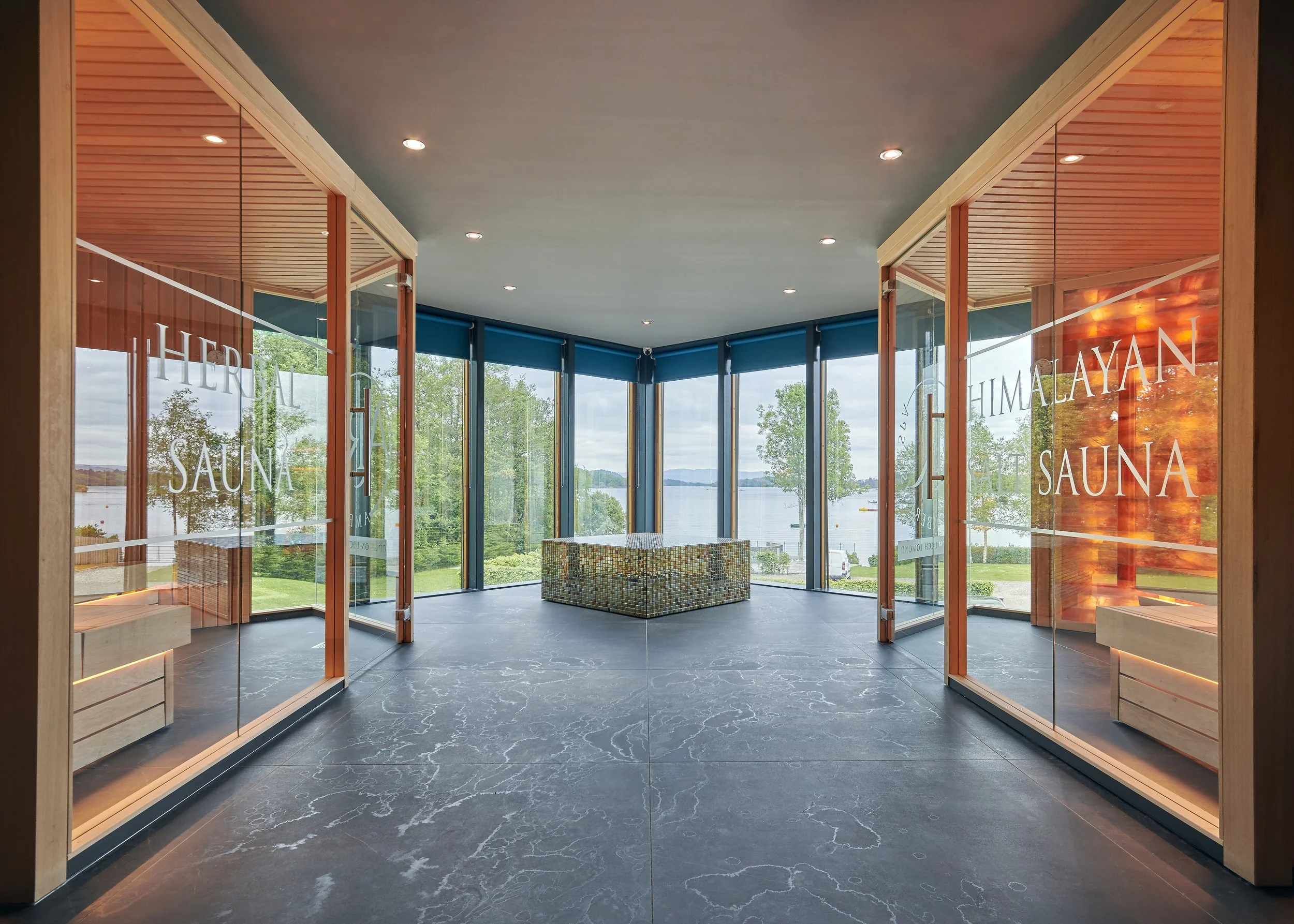 Modern spa interior with glass saunas and panoramic lake view, featuring herbal and Himalayan salt sauna rooms.