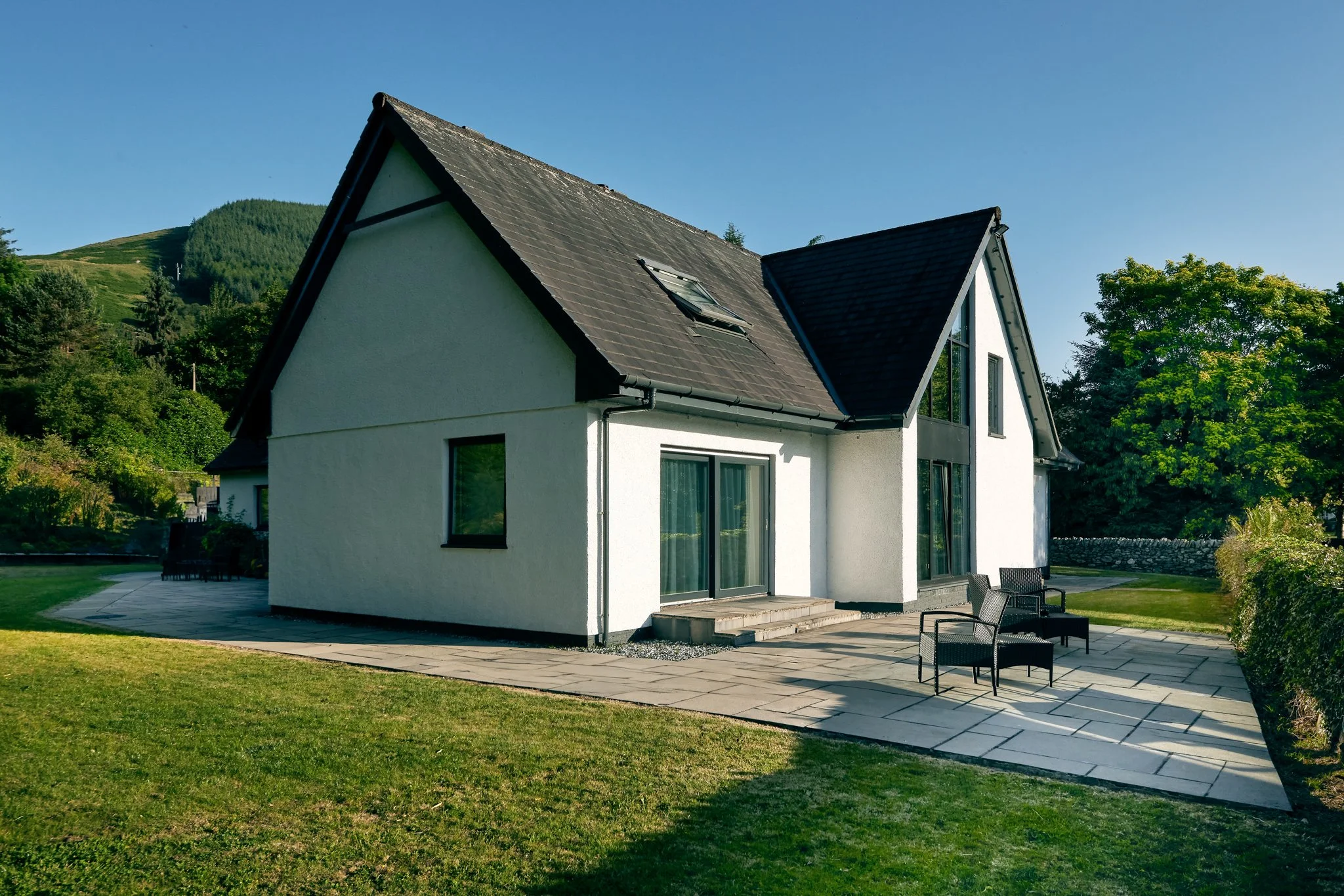 Modern white detached house with dark pitched roof and garden patio, photographed in bright afternoon light in the Scottish countryside.