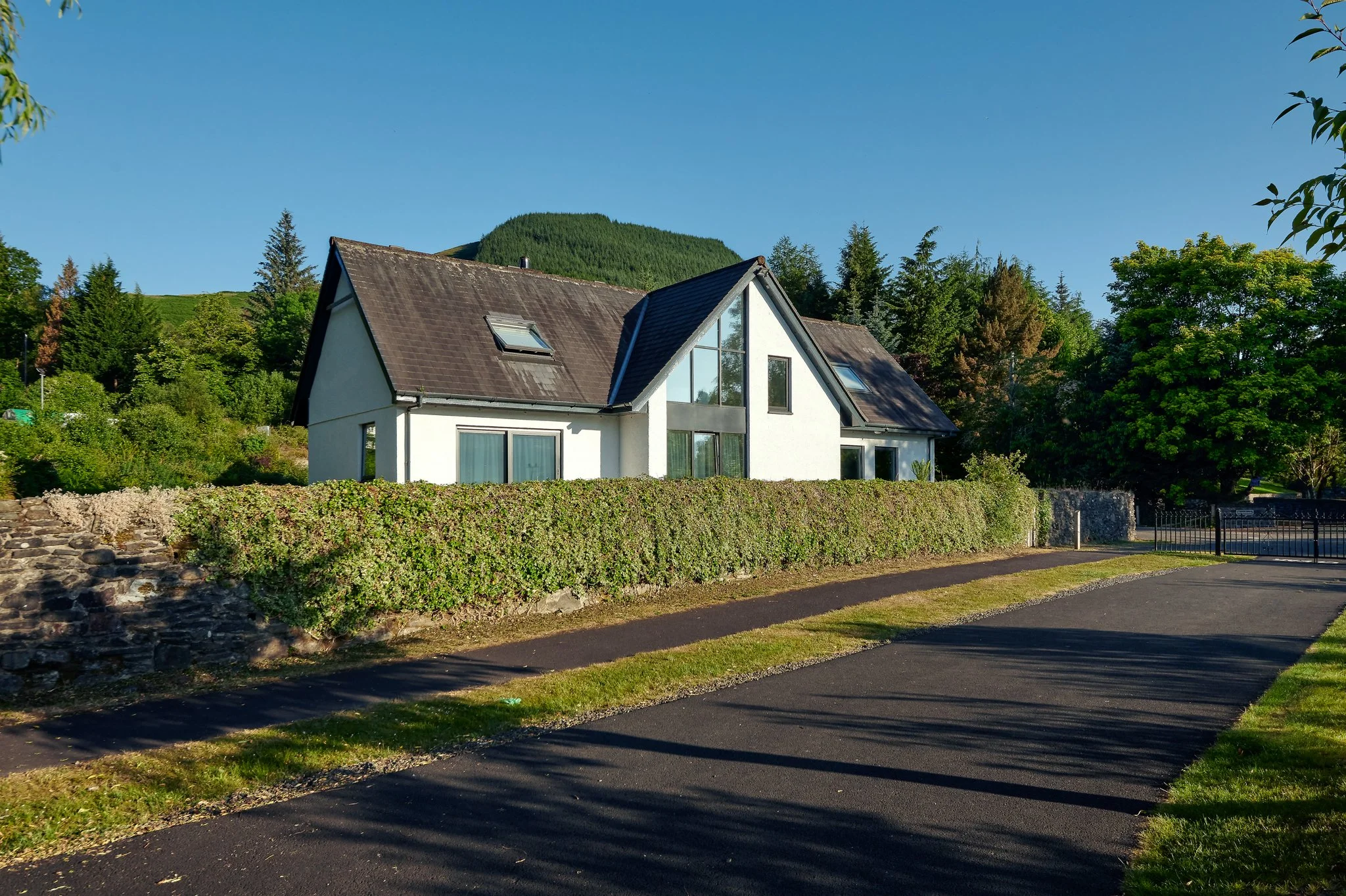 Contemporary white house with large windows and dark roof, set against a lush green hillside and surrounded by tall trees and hedges in rural Scotland.