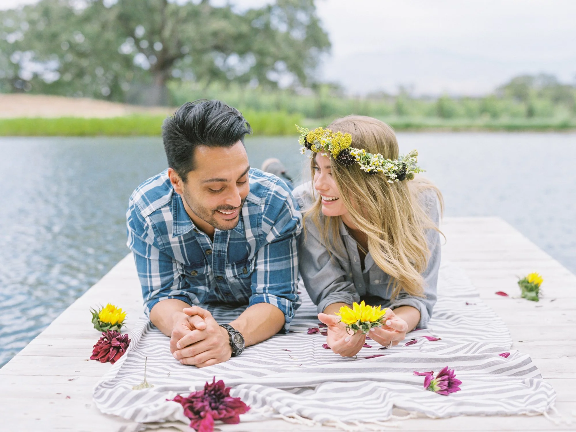 Couple lying on a wooden dock by the pond at Refugio Ranch, smiling at each other, surrounded by flowers and greenery.